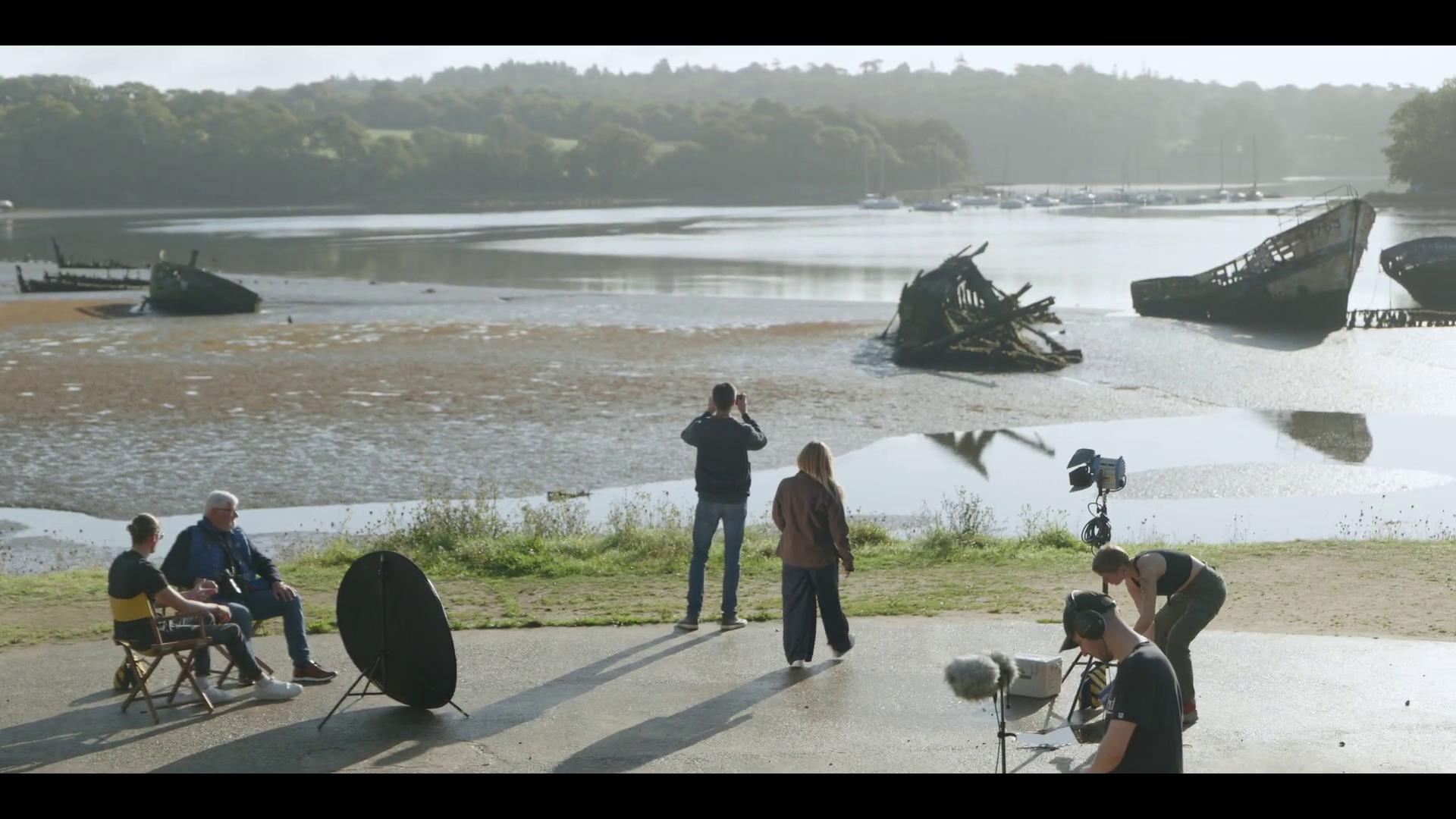 Une équipe de production réalise une séance photo ou un tournage en extérieur. Au premier plan, plusieurs personnes, certains avec du matériel comme des réflecteurs et une caméra, sont en train de travailler. À l'arrière-plan, une rivière avec des embarcations échouées ou en décomposition, et au loin, des bateaux amarrés sur la rivière. Plusieurs personnes sont en train de prendre des photos ou de filmer.