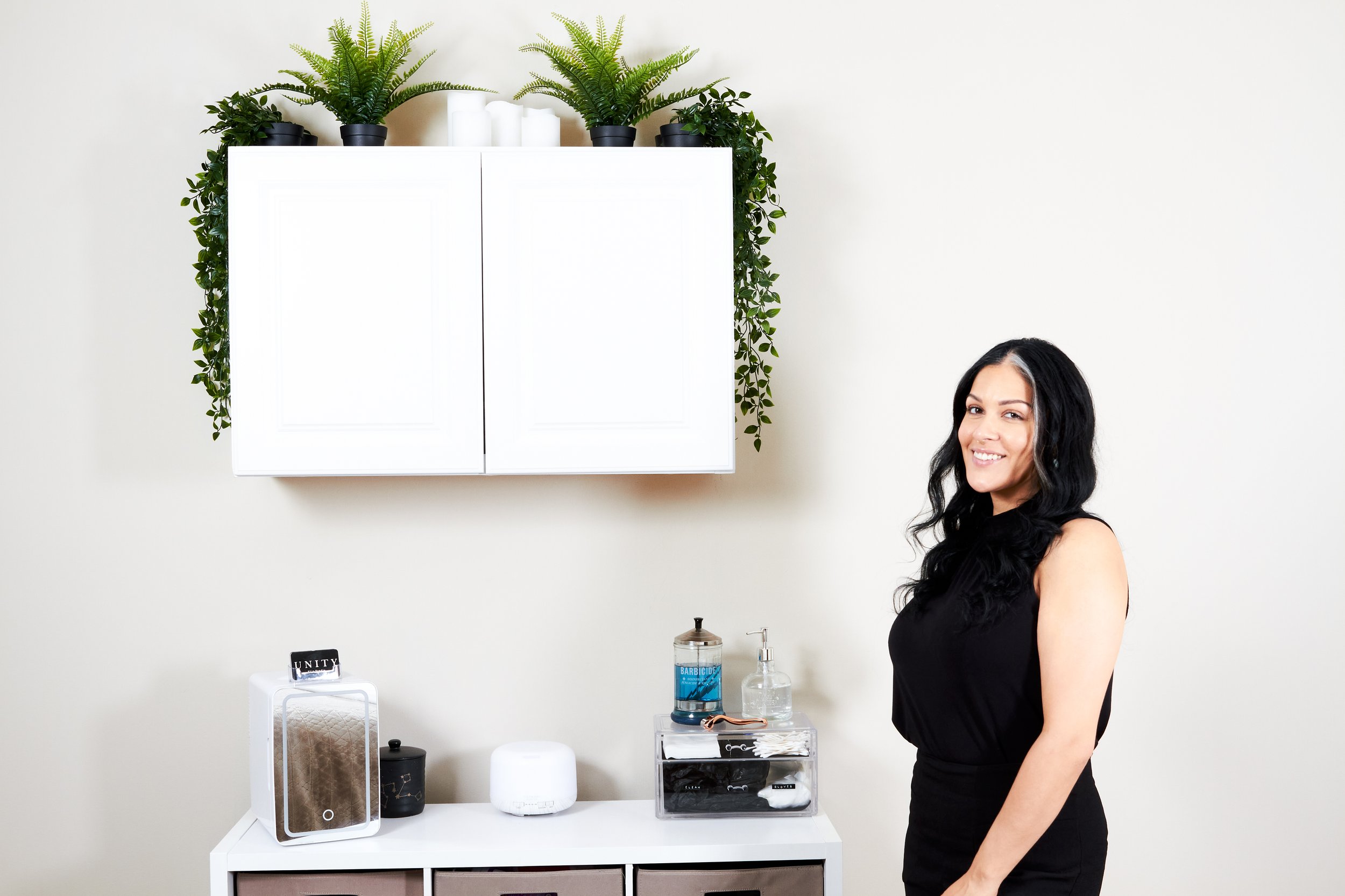 Woman smiling in a minimalistic room, with a white cabinet, plants, and various personal care items.