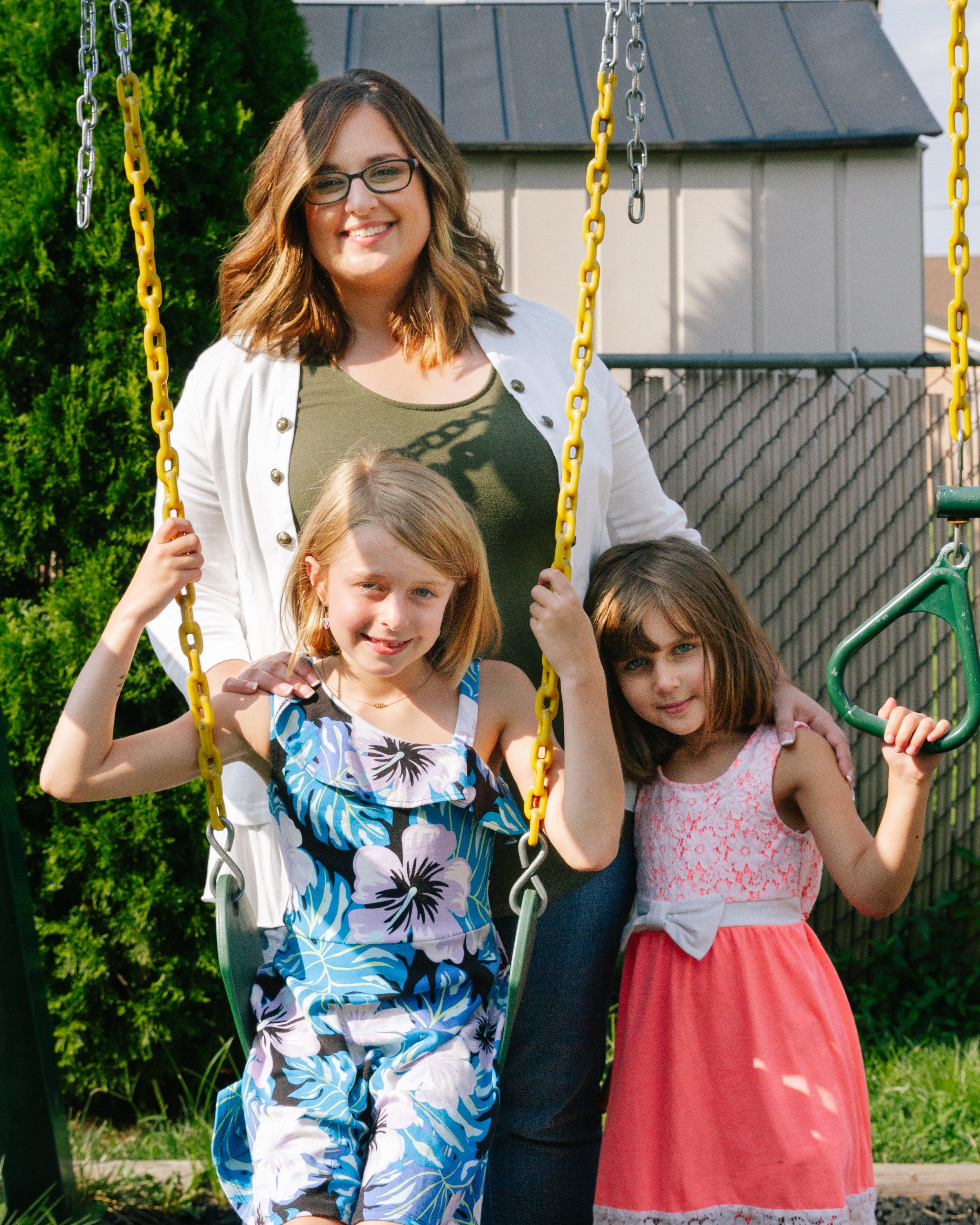 A woman stands behind two young girls on swings in a playground, smiling. One girl wears a floral dress, the other a pink dress.