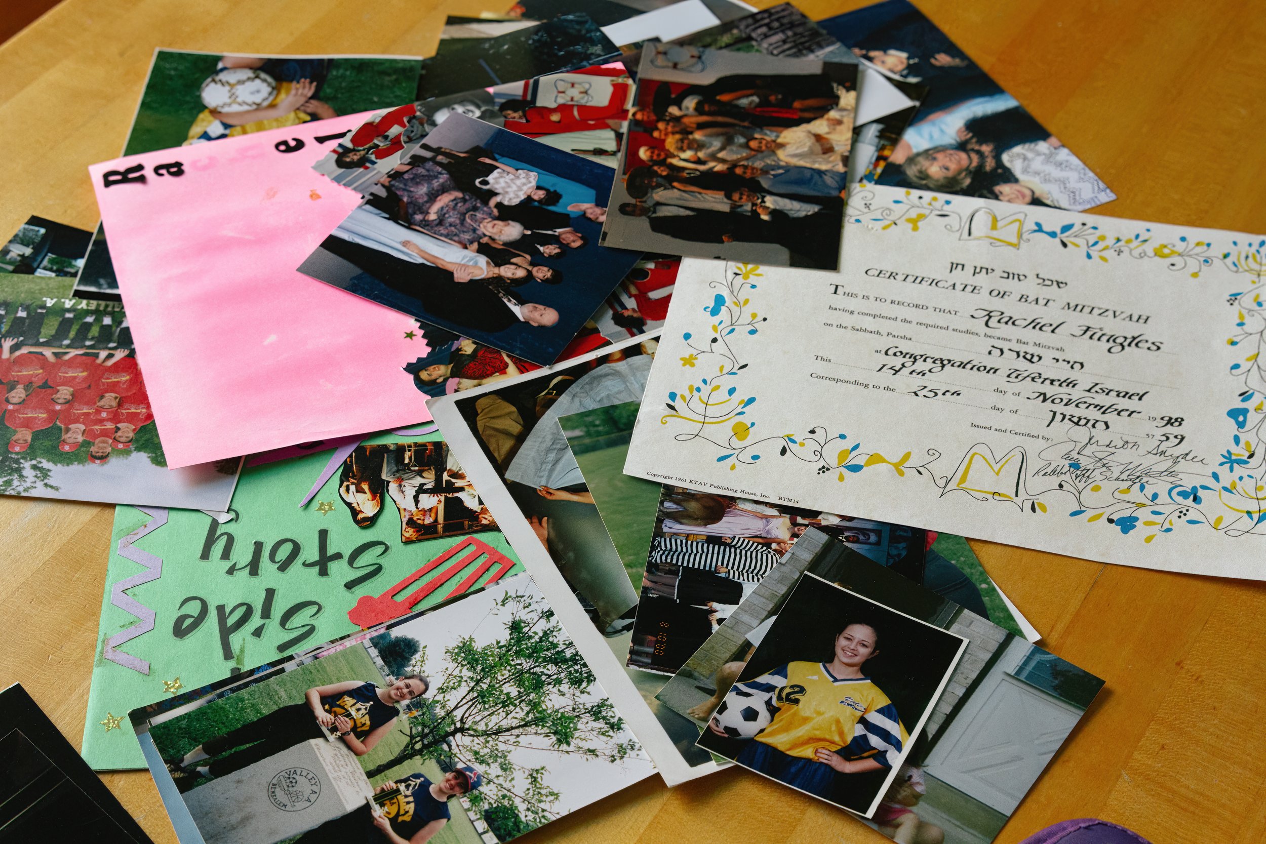 A collection of photographs and a Bat Mitzvah certificate scattered on a wooden table. One prominent photo shows a girl in a yellow soccer jersey holding a soccer ball. The certificate is decorated with floral designs and includes handwritten details