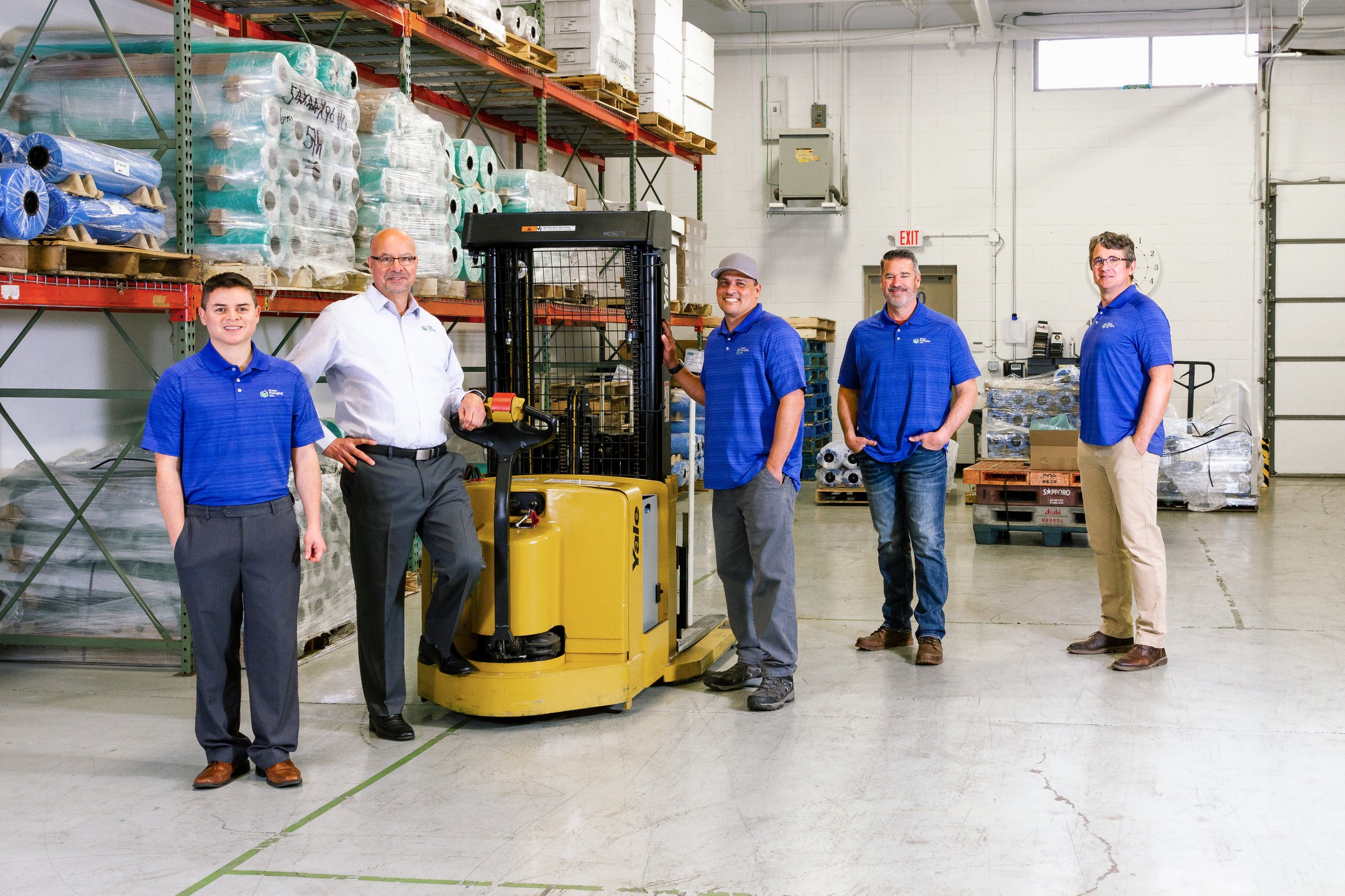 Five men standing in a warehouse with boxed and rolled products, next to a yellow warehouse forklift.