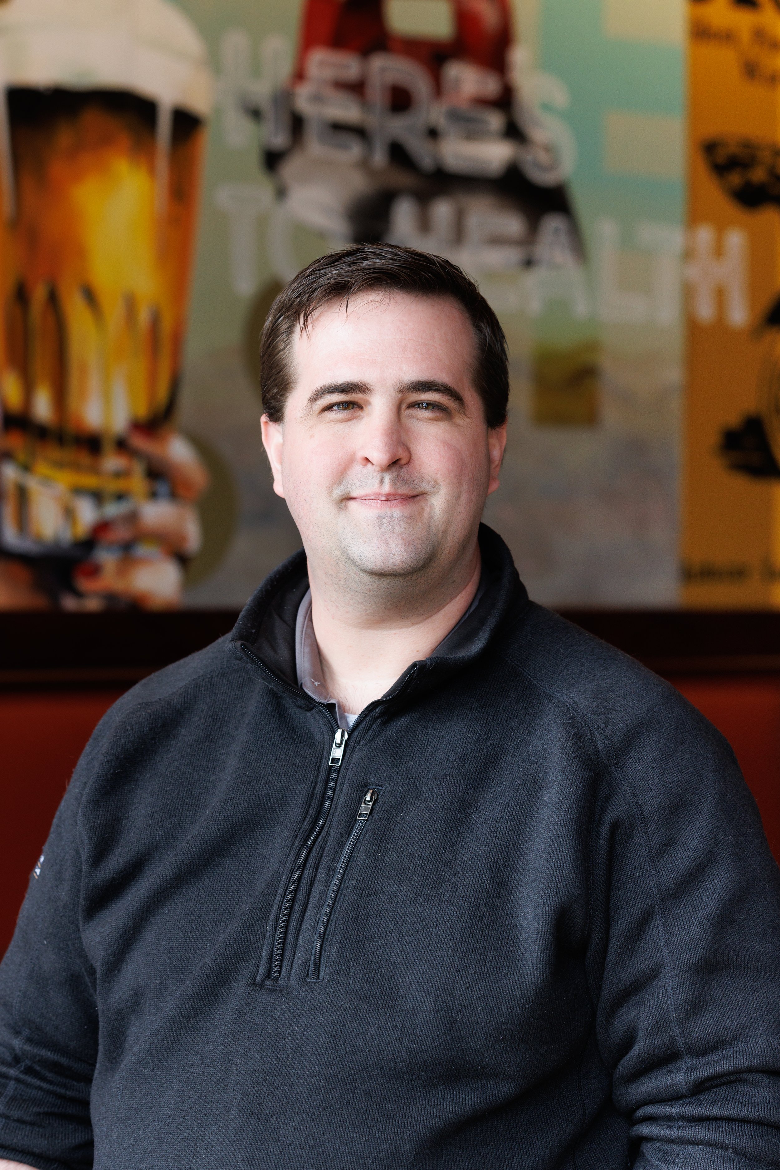 Man in black zip-up sweater smiling with a blurred background of a bar or pub setting.