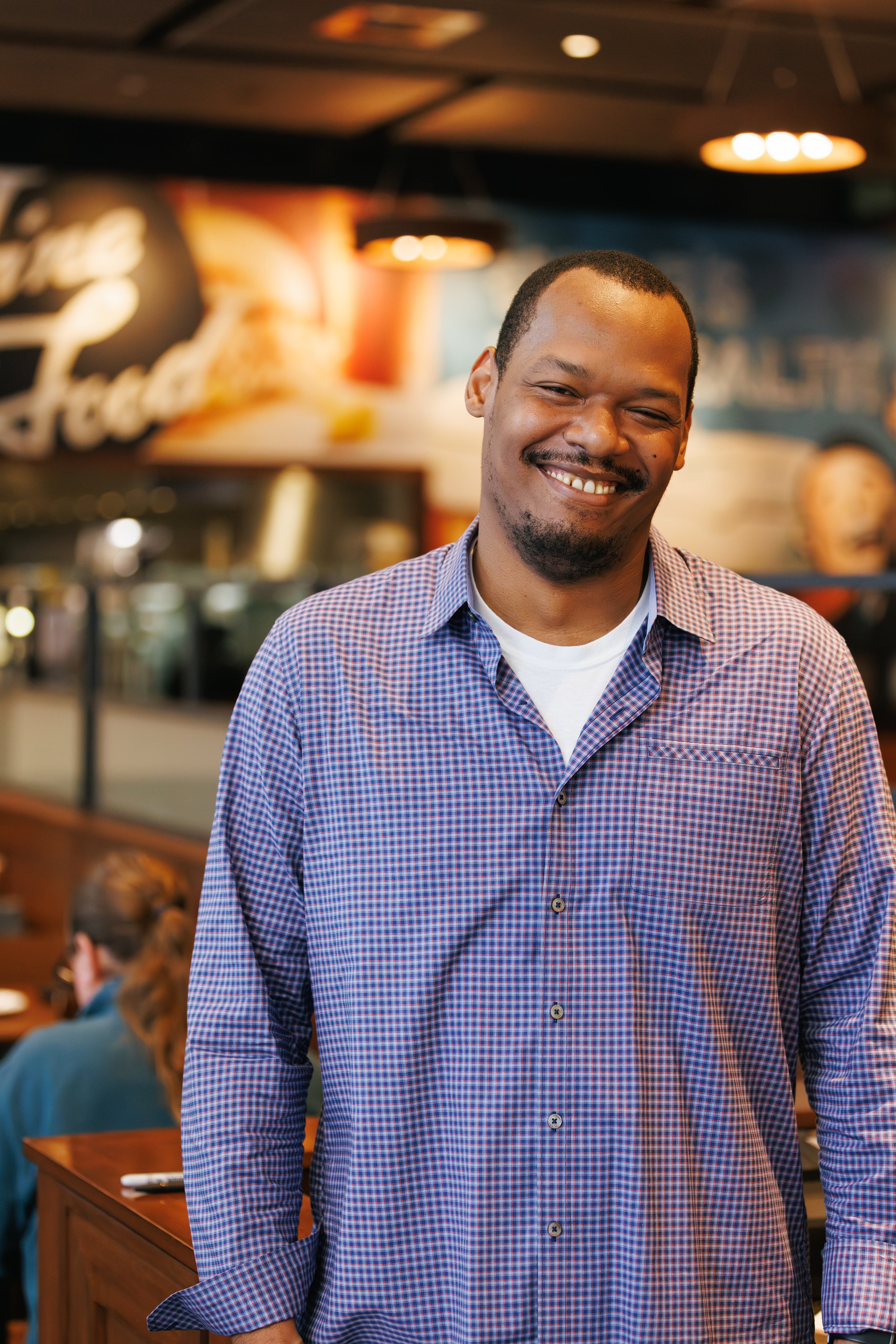 Man smiling in a casual restaurant wearing a blue checkered shirt