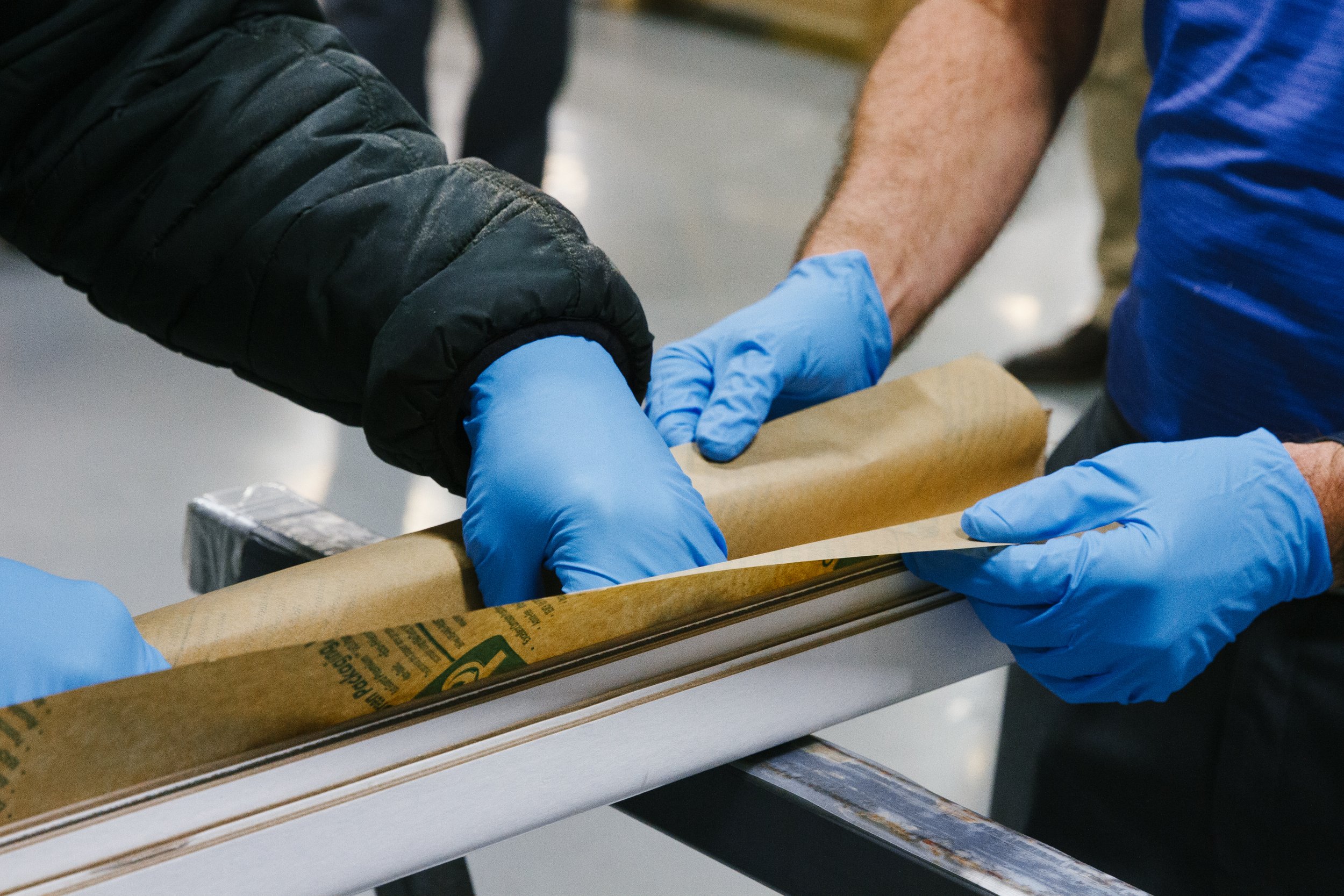 Two people wearing blue gloves handling brown paper over a metal surface.