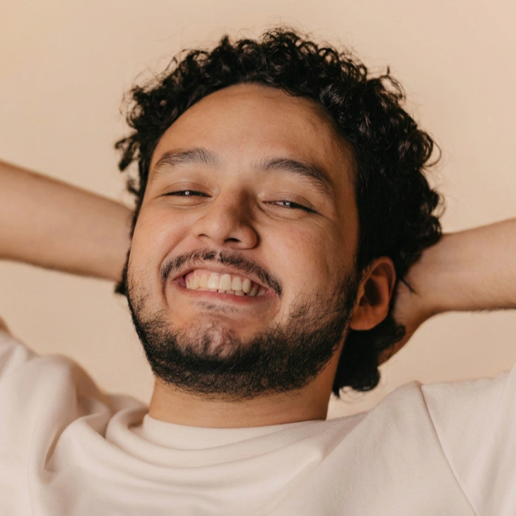 Young man with curly hair smiling and relaxing with arms behind head.