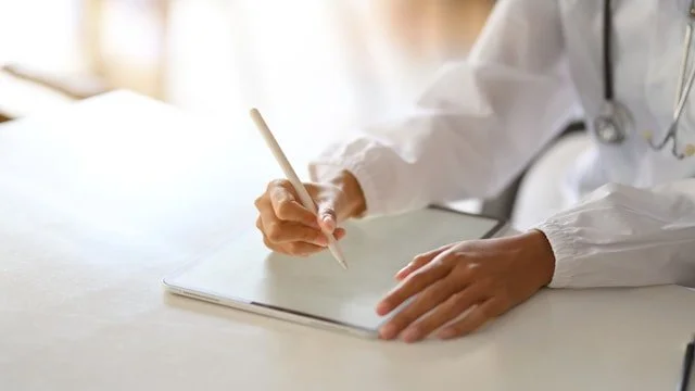 A person wearing a white coat and stethoscope writing on a notepad at a desk.