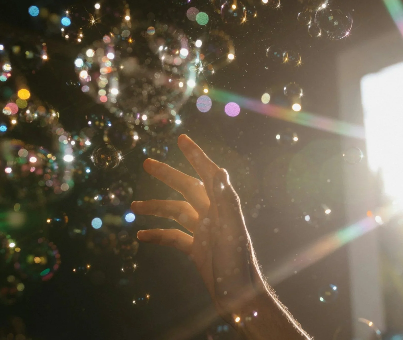 A person's hand reaching towards a decorated Christmas tree with colorful lights, ornaments, and a bright light source on the right side of the image, creating a glow and a festive atmosphere.
