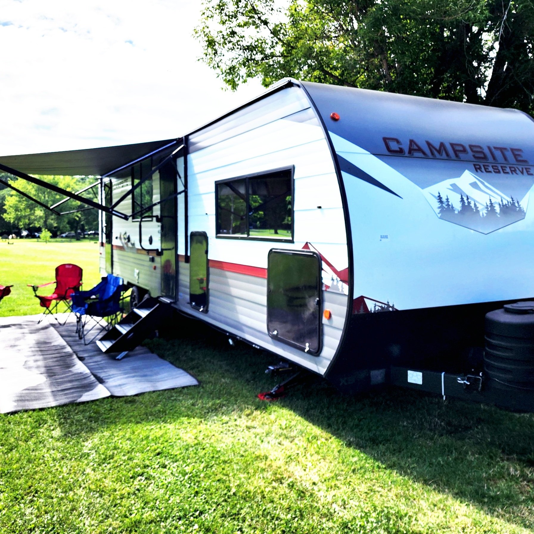 Travel trailer parked on grass with awning extended, red and blue camp chairs, and a logo that reads 'Campsite Reserve.'