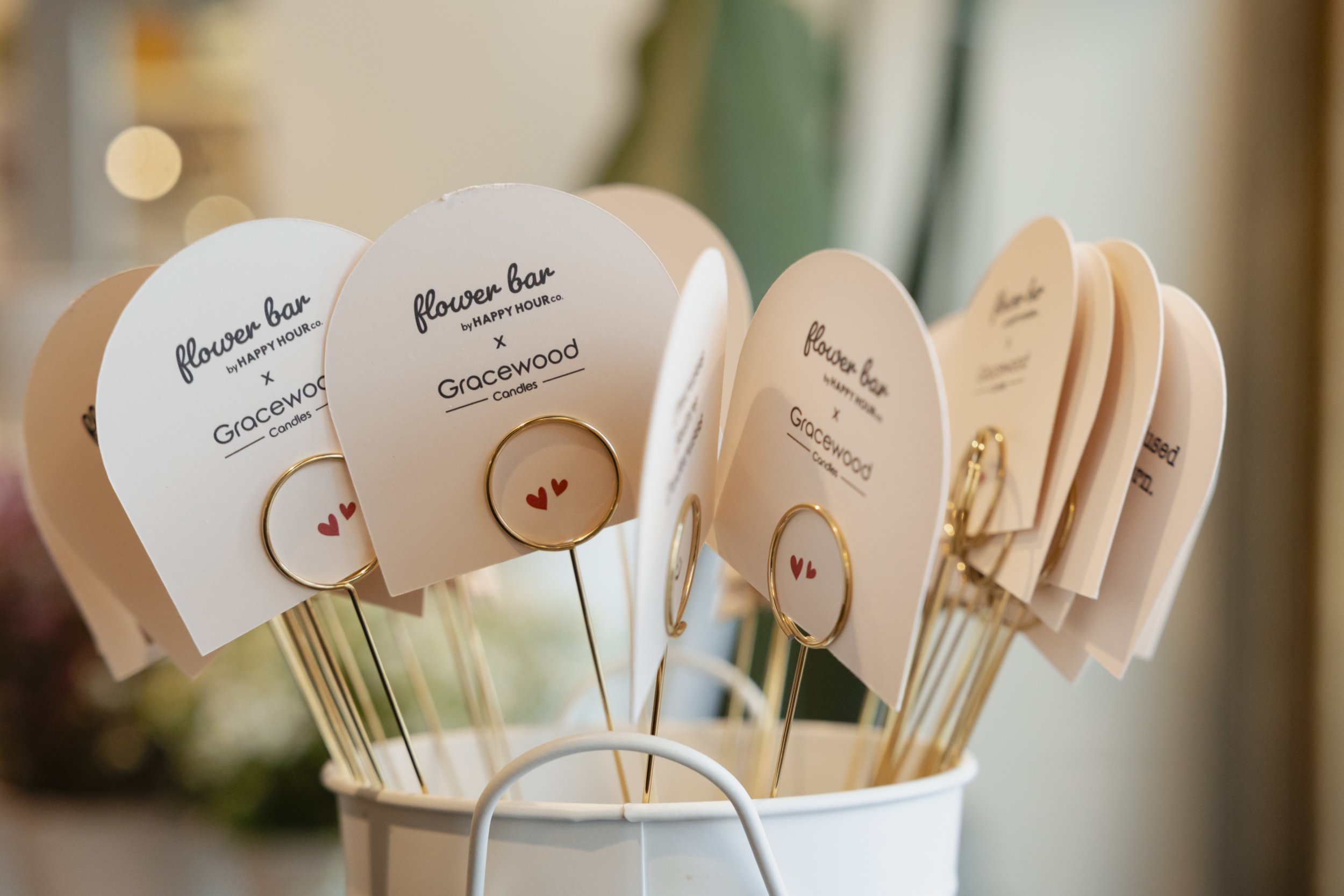 Display of small white cards with black text for a flower bar and candle display, attached to gold wire and decorated with small red hearts, in a white container.