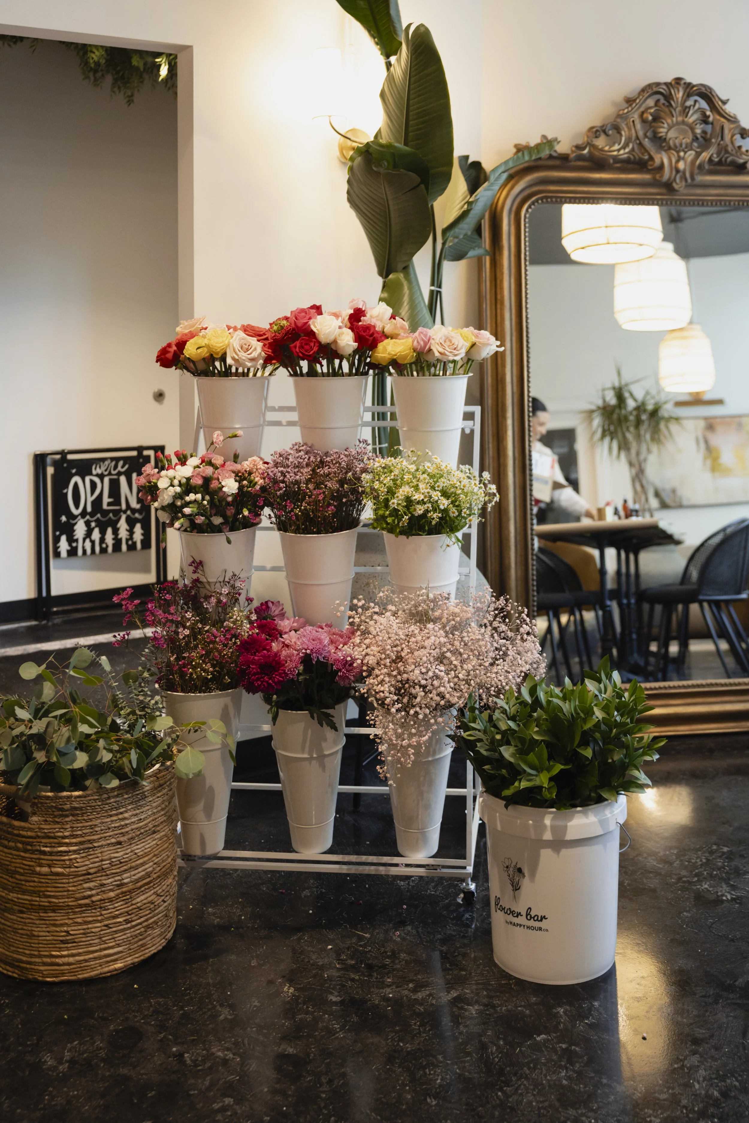 A display of various colorful flowers in white flower pots on a metal stand, next to a large ornate mirror and a woven basket, in a cozy indoor setting with hanging lights.