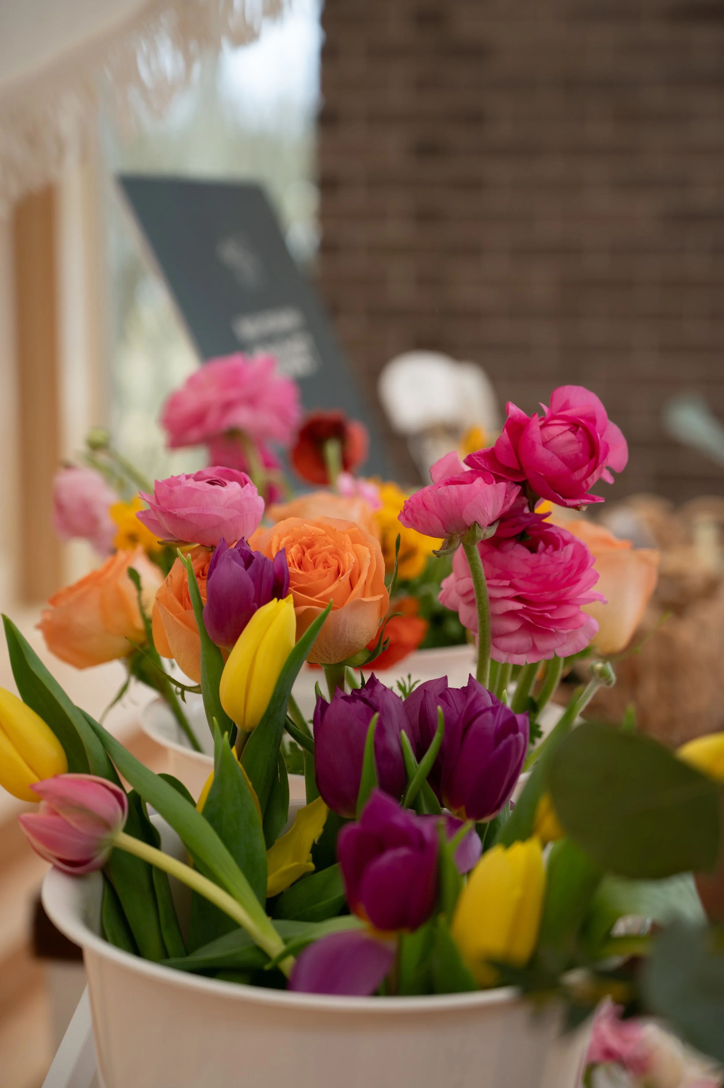 A colorful bouquet of tulips and roses in a white vase, with blurred background including a blackboard and some decorative items.