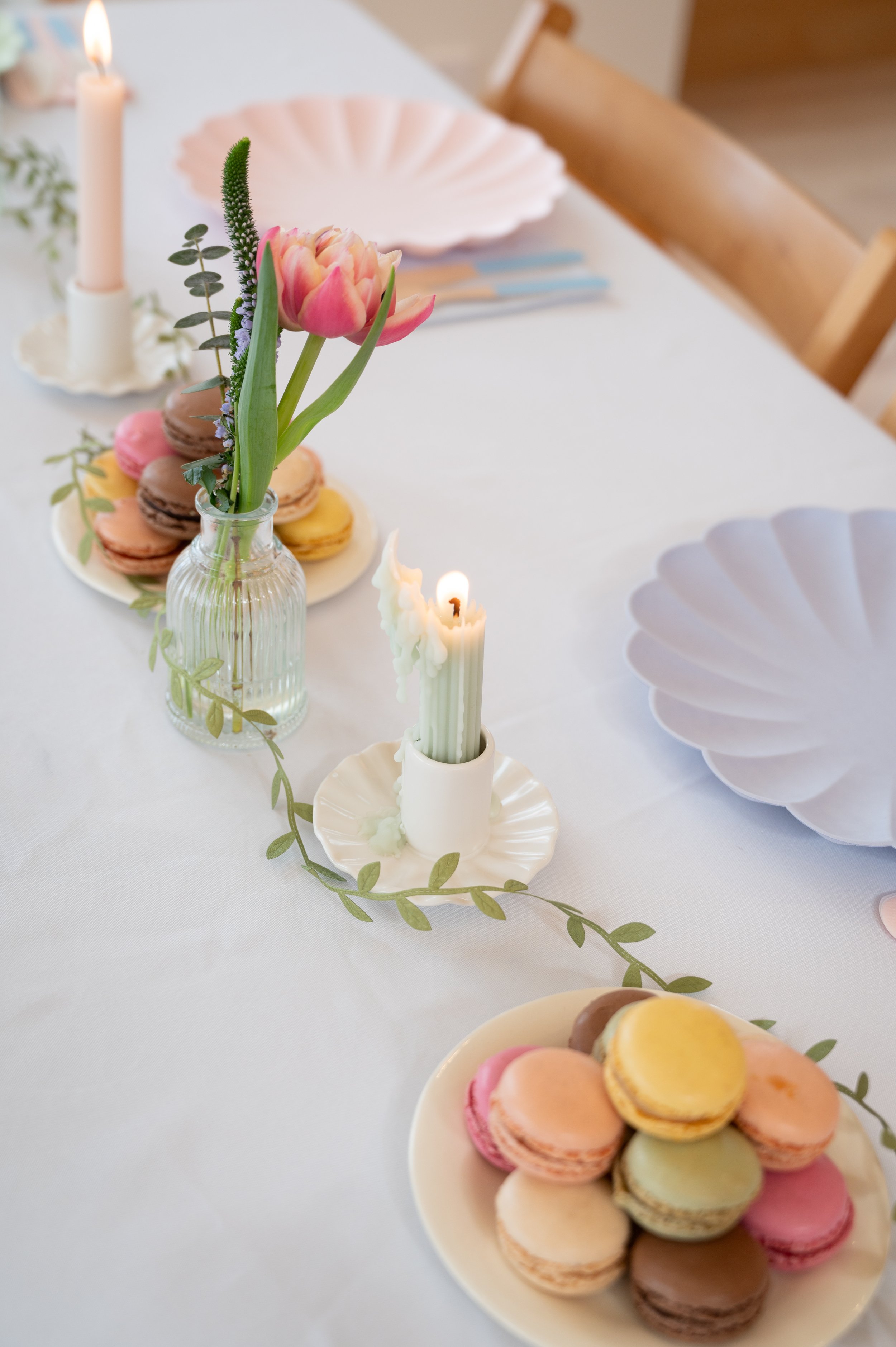 Decorative table setting with a pink tulip in a glass vase, colorful macarons on cake plates, and lit candles, with pastel-colored plates and a white tablecloth.
