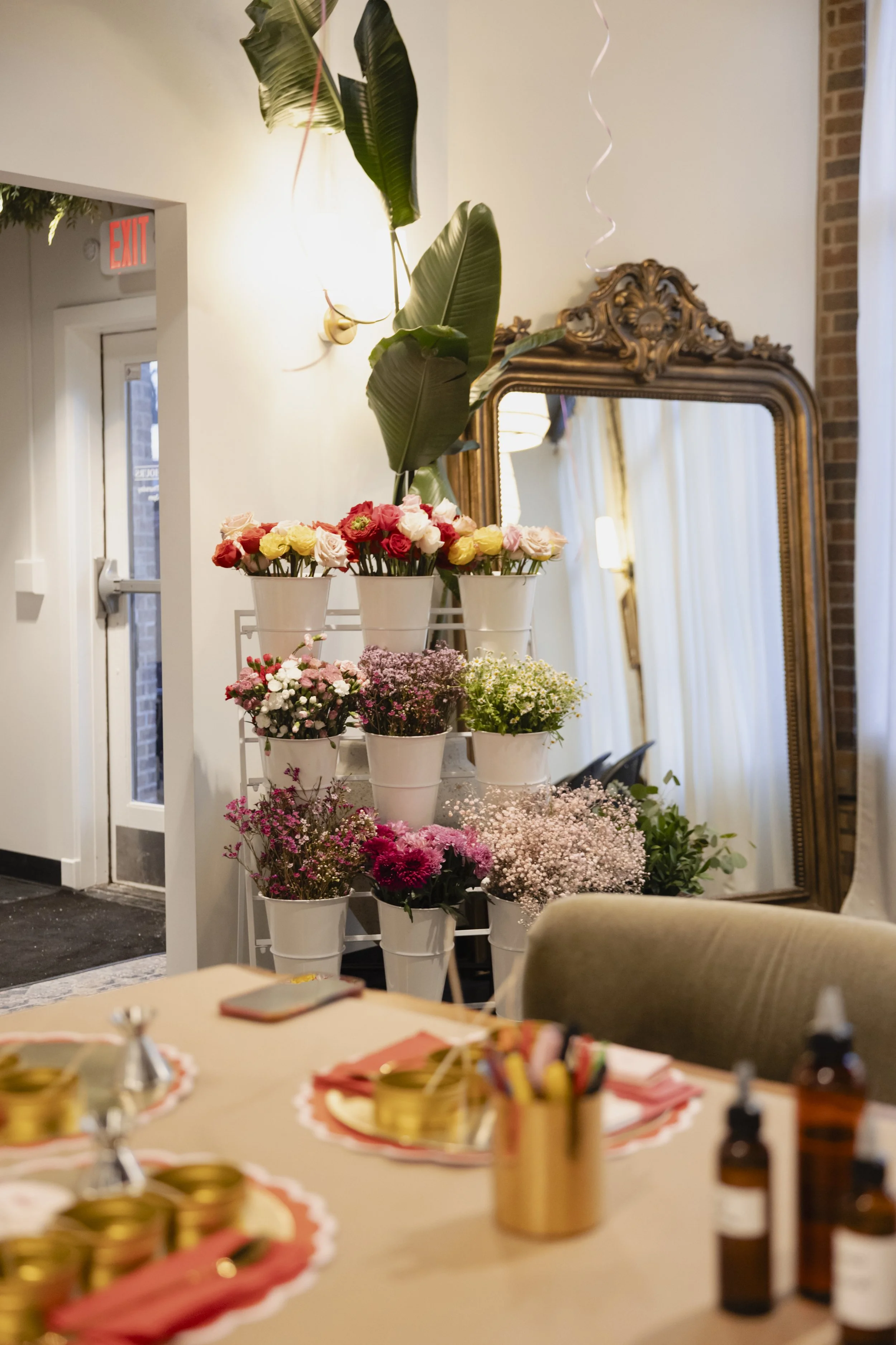 Flower arrangement with pink, yellow, and white roses, and pink baby's breath in white pots on a metal stand, reflected in an ornate gold-framed mirror, with green leaves and white curtains in the background.