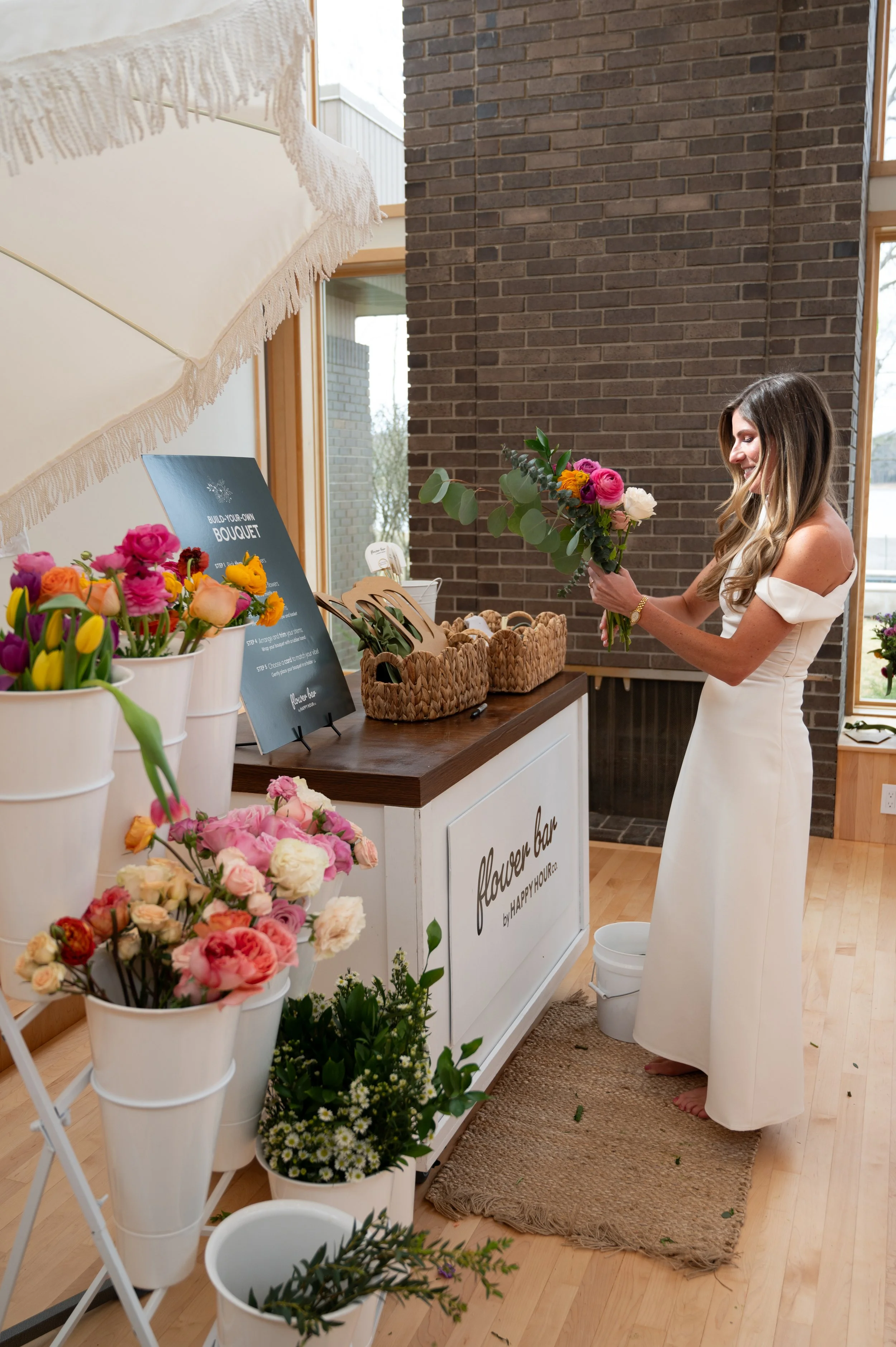 A woman in a white dress arranging a bouquet of pink, white, and orange flowers at a flower shop interior with flowers in white buckets and a sign that reads ‘flower bar’.