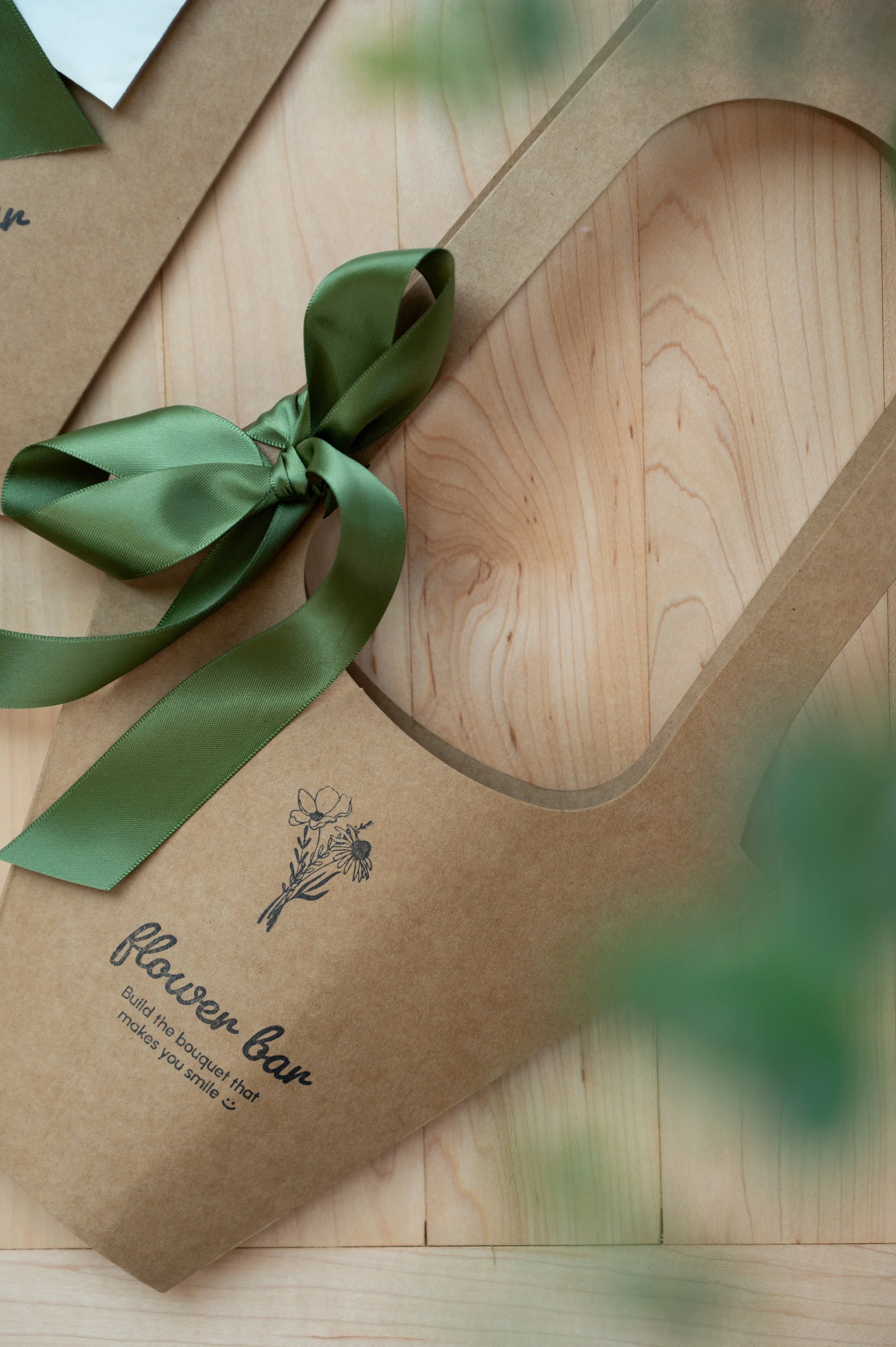 A brown paper bouquet holder with a green satin ribbon tied in a bow, labeled 'flower bar' with a small flower illustration and the phrase 'Build the bouquet that makes you smile',' placed on a wooden surface.