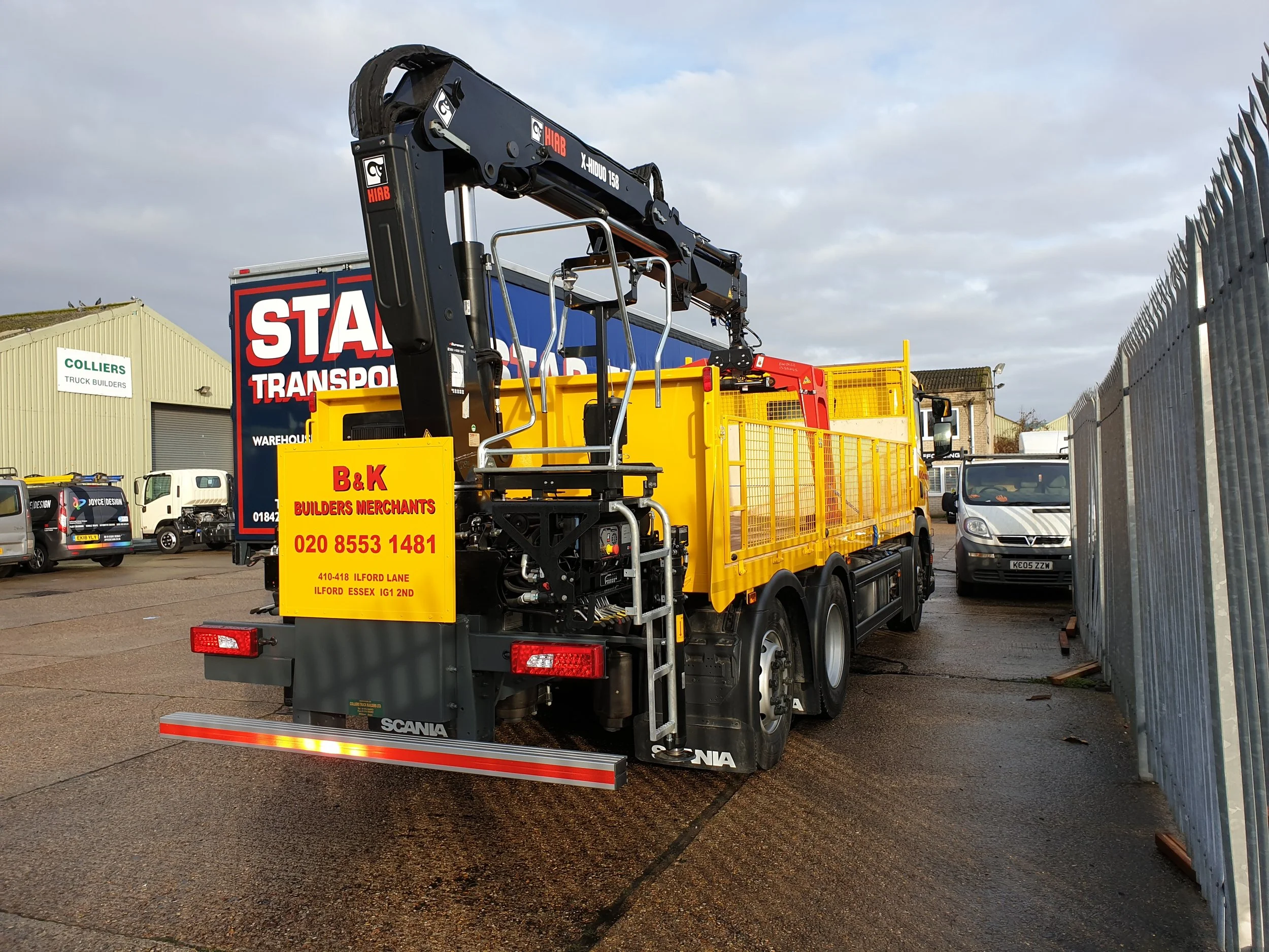 Yellow and black work truck with a crane arm, parked in lot with other vehicles and a green warehouse in background.