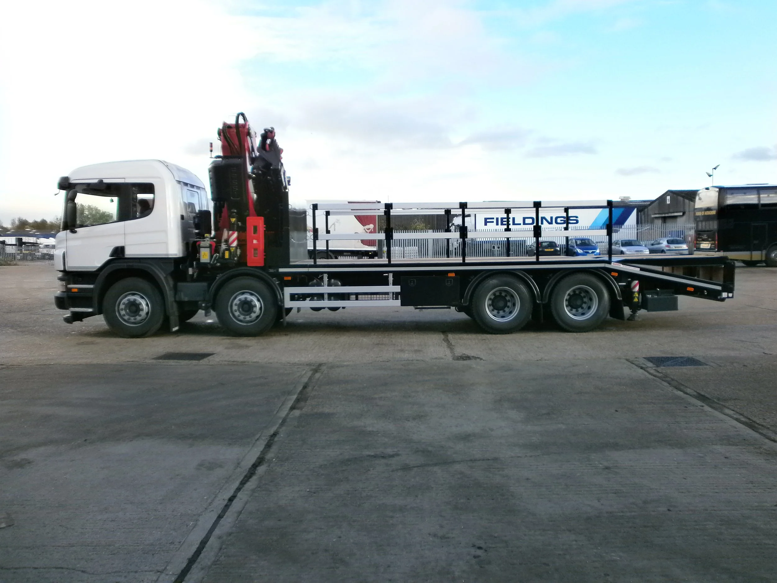A flatbed tow truck with a crane parked in an open lot, with a few cars and a large building labeled 'FIELDINGS' in the background.