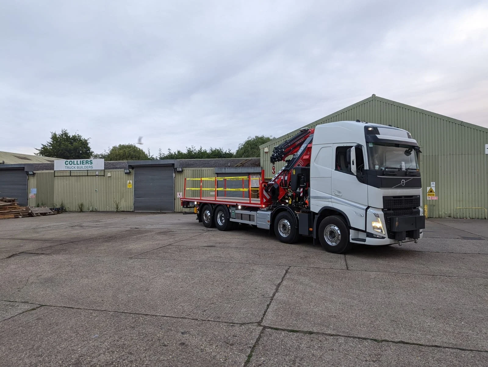 A white truck with a hydraulic crane parked in front of a green industrial building with a sign that reads 'COLLIERS TRUCK BUILDERS'.