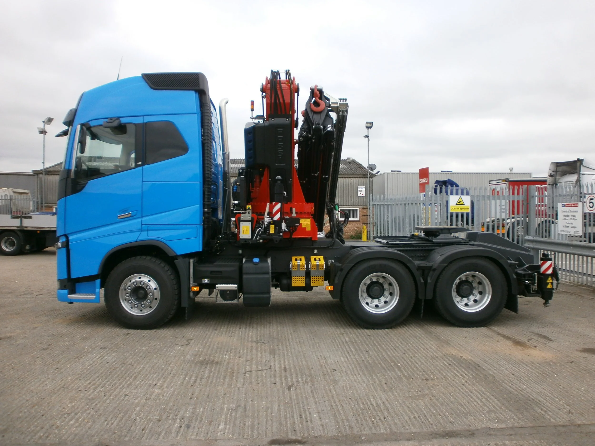 Blue truck with a mounted crane in a parking lot surrounded by metal fences and other trucks.