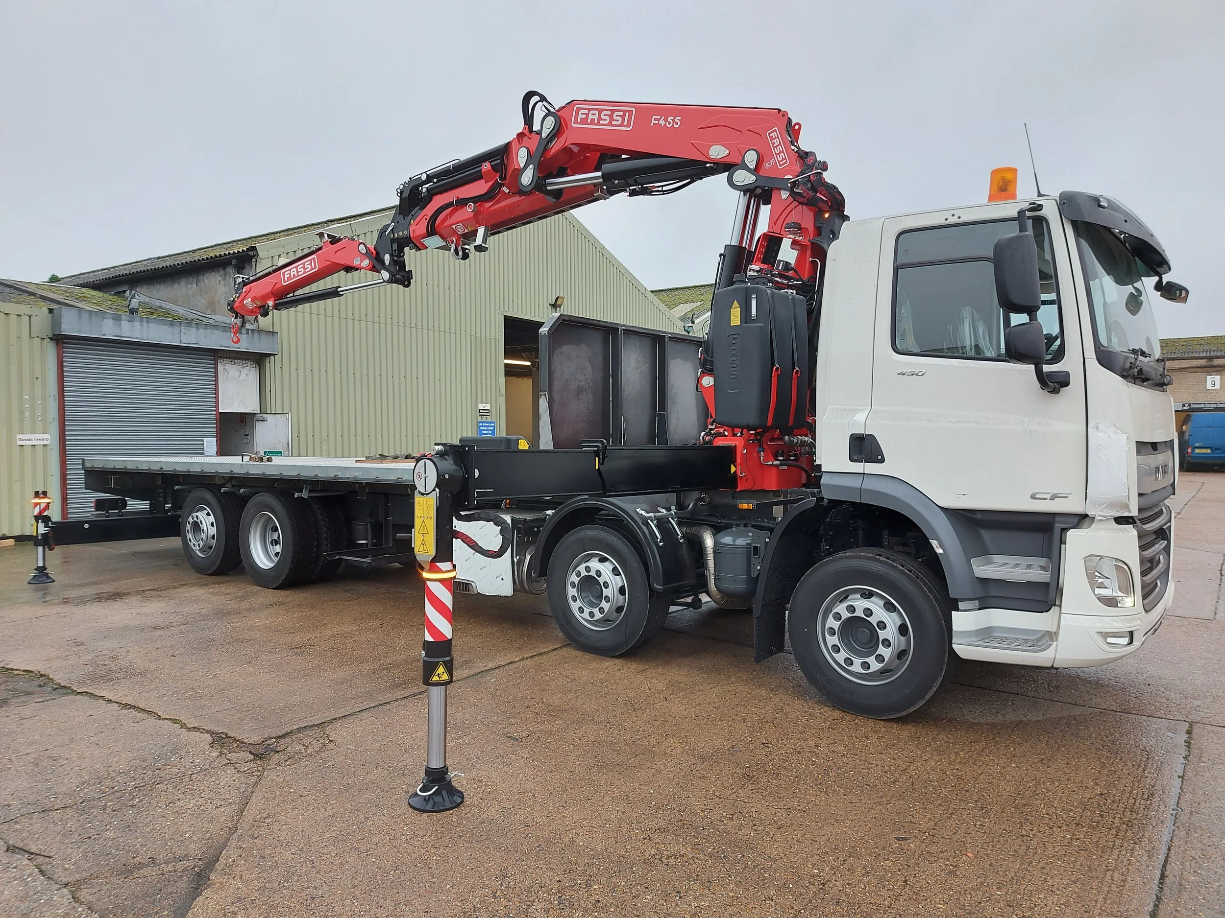 A flatbed truck with a mounted red hydraulic crane in an industrial yard.