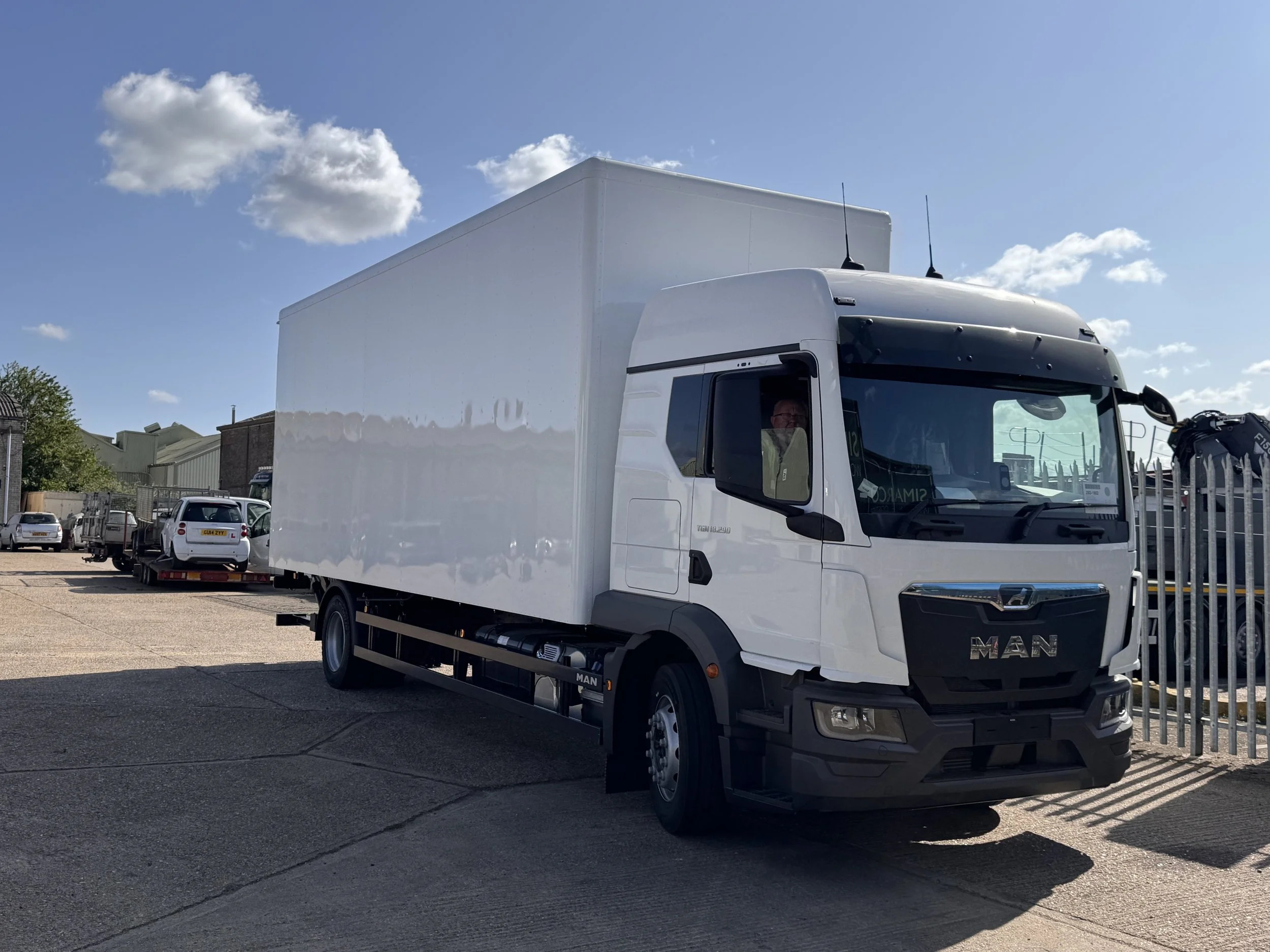 White box truck parked in an outdoor lot with other vehicles and buildings, under a blue sky with clouds.