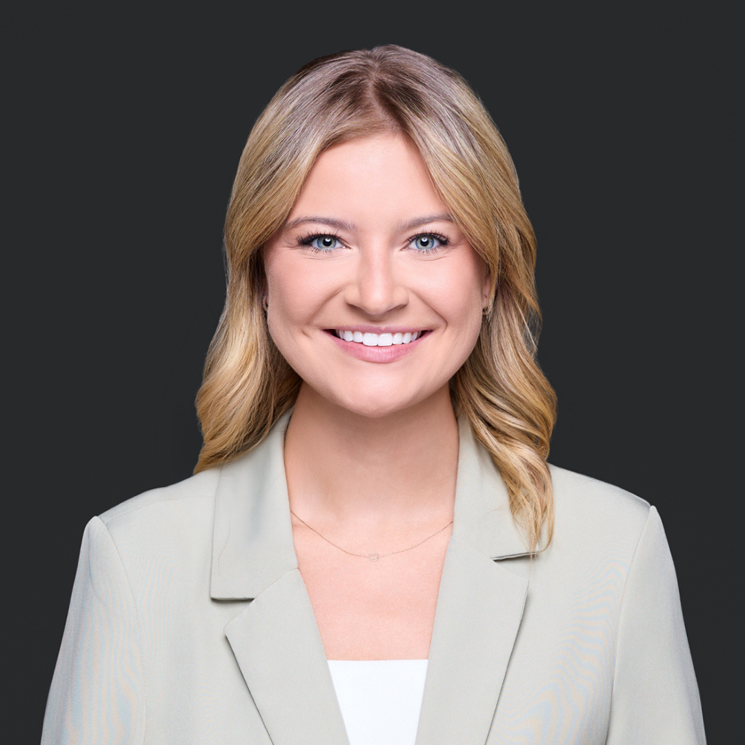 Close-up portrait of a Red Deer insurance woman with shoulder-length wavy blonde hair, smiling, wearing a tan. blazer, against a dark gray background.