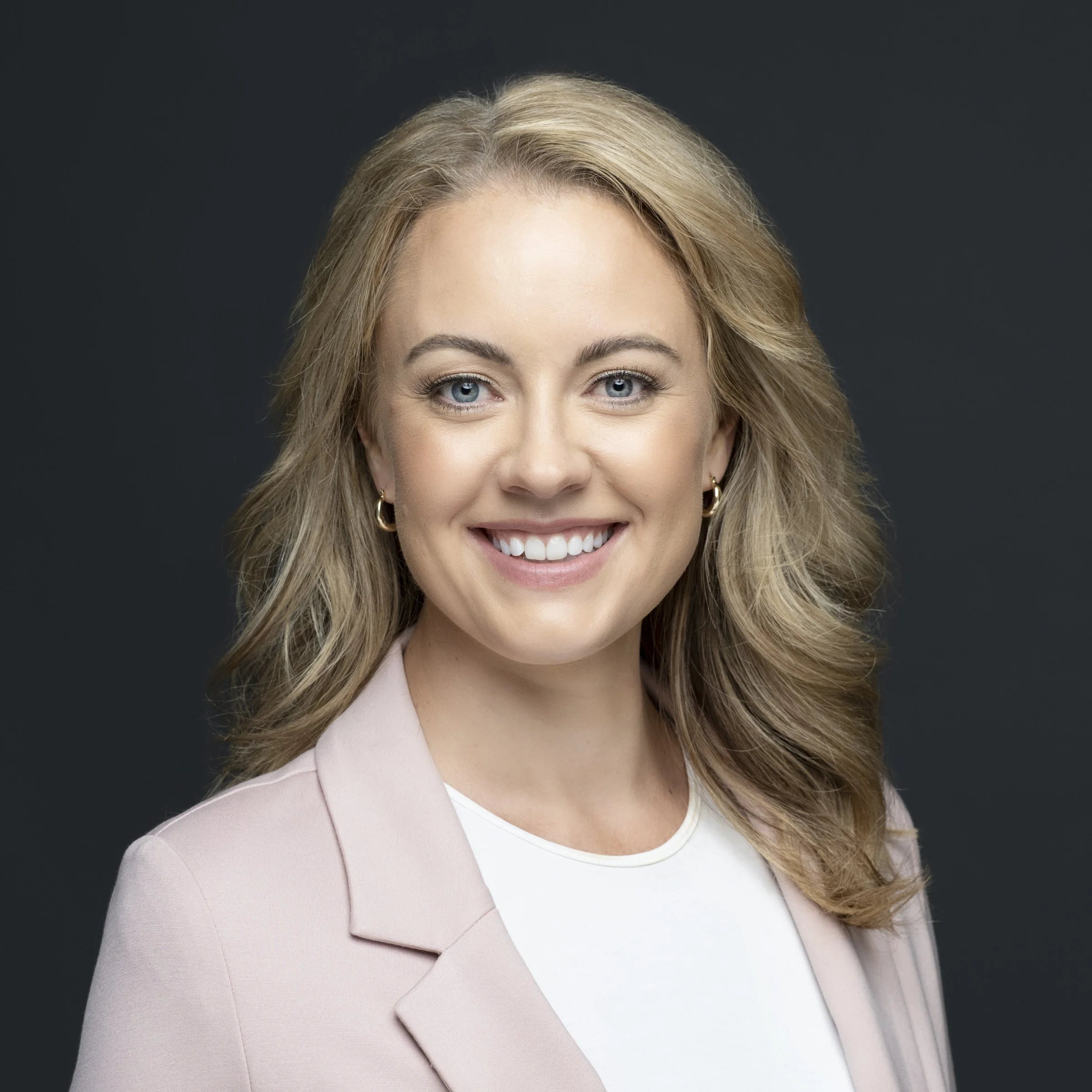 Headshot of a smiling woman with wavy blonde hair, blue eyes, wearing a light pink blazer and white top against a dark background.