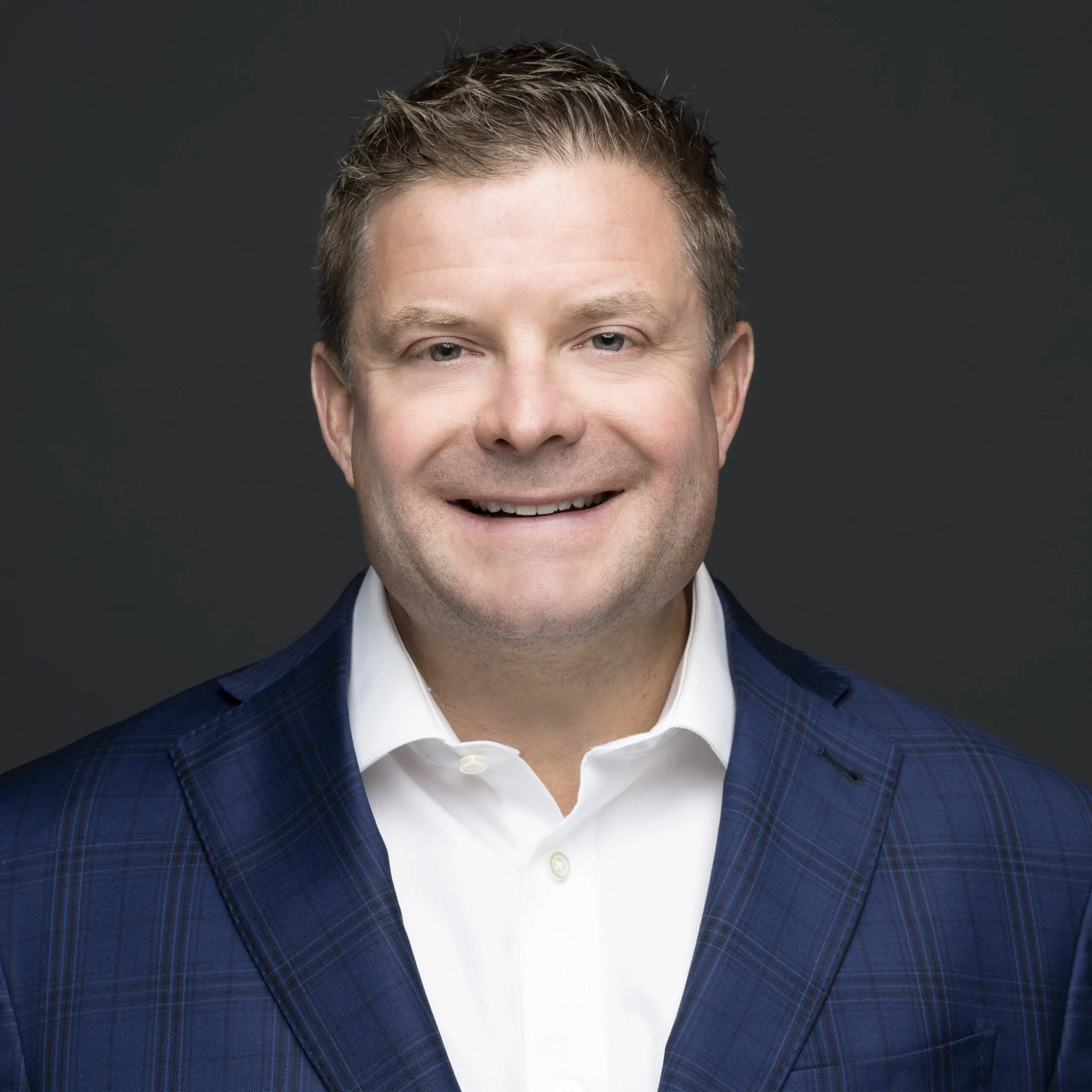 A professional headshot of a man wearing a white shirt and a blue plaid blazer, smiling against a dark gray background.