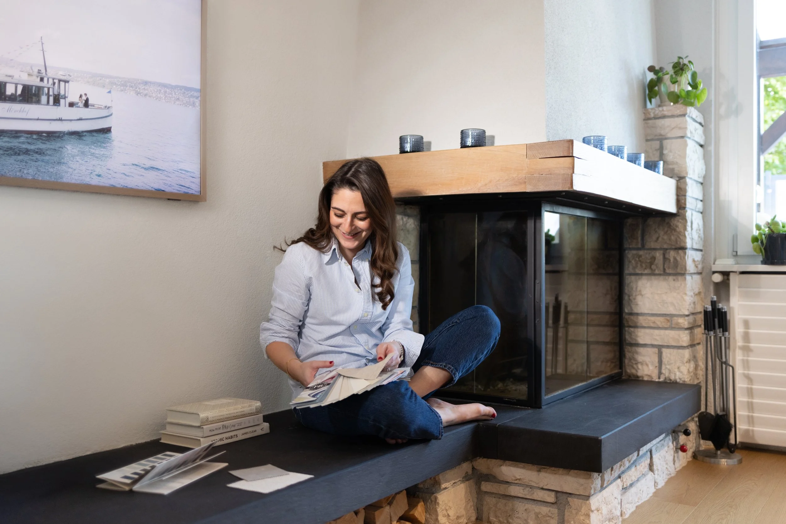 A woman sitting on a black ledge in front of a stone fireplace, looking through a fan deck of paint samples and smiling. There are books and paint swatches on the ledge, with a large window to her right. Cosy fireplace tv room, snug, nook room fire