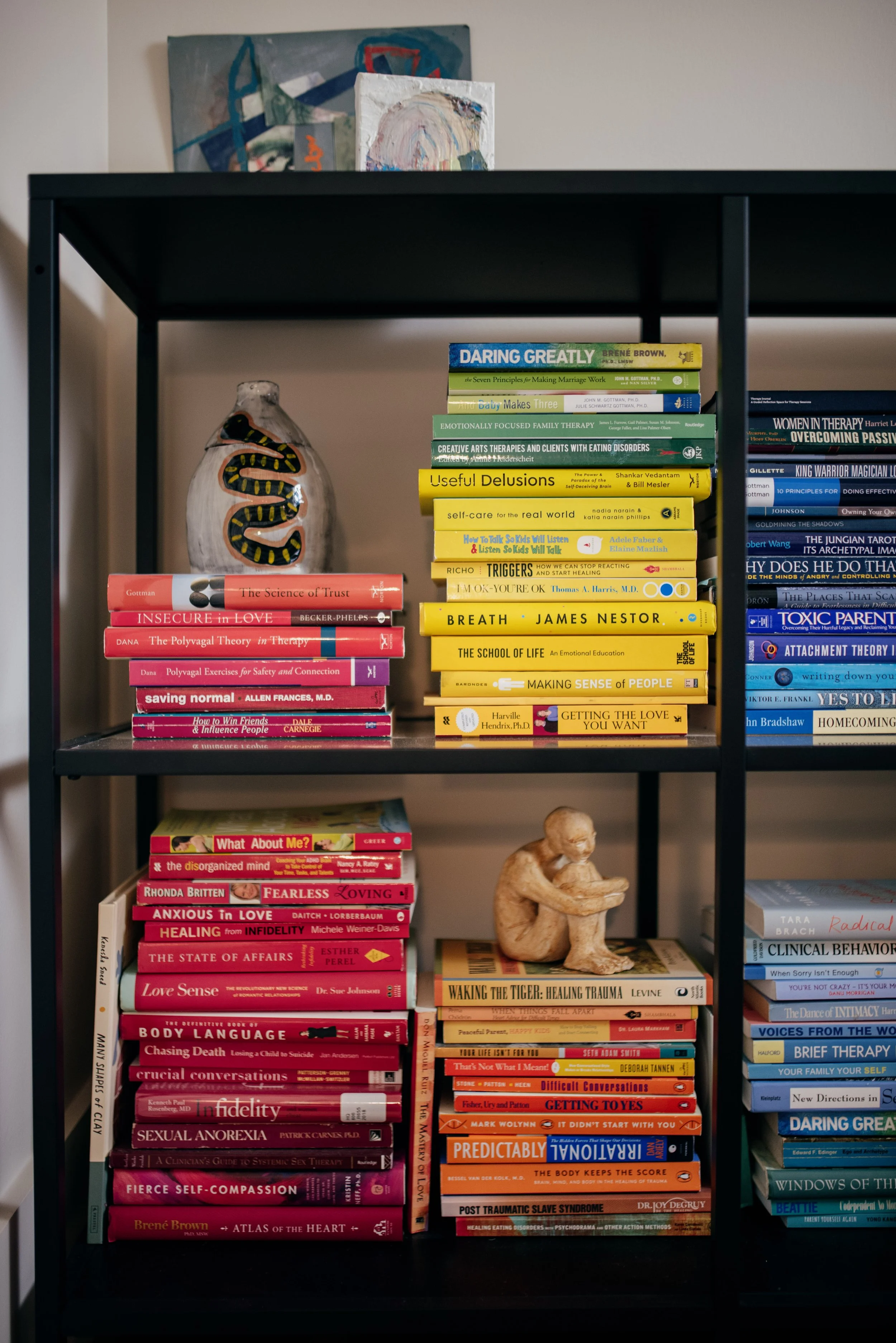 Bookshelf containing multiple stacks of books on psychology, therapy, and self-help topics, a ceramic snake, and a small sculpture of a person sitting and hugging their knees.