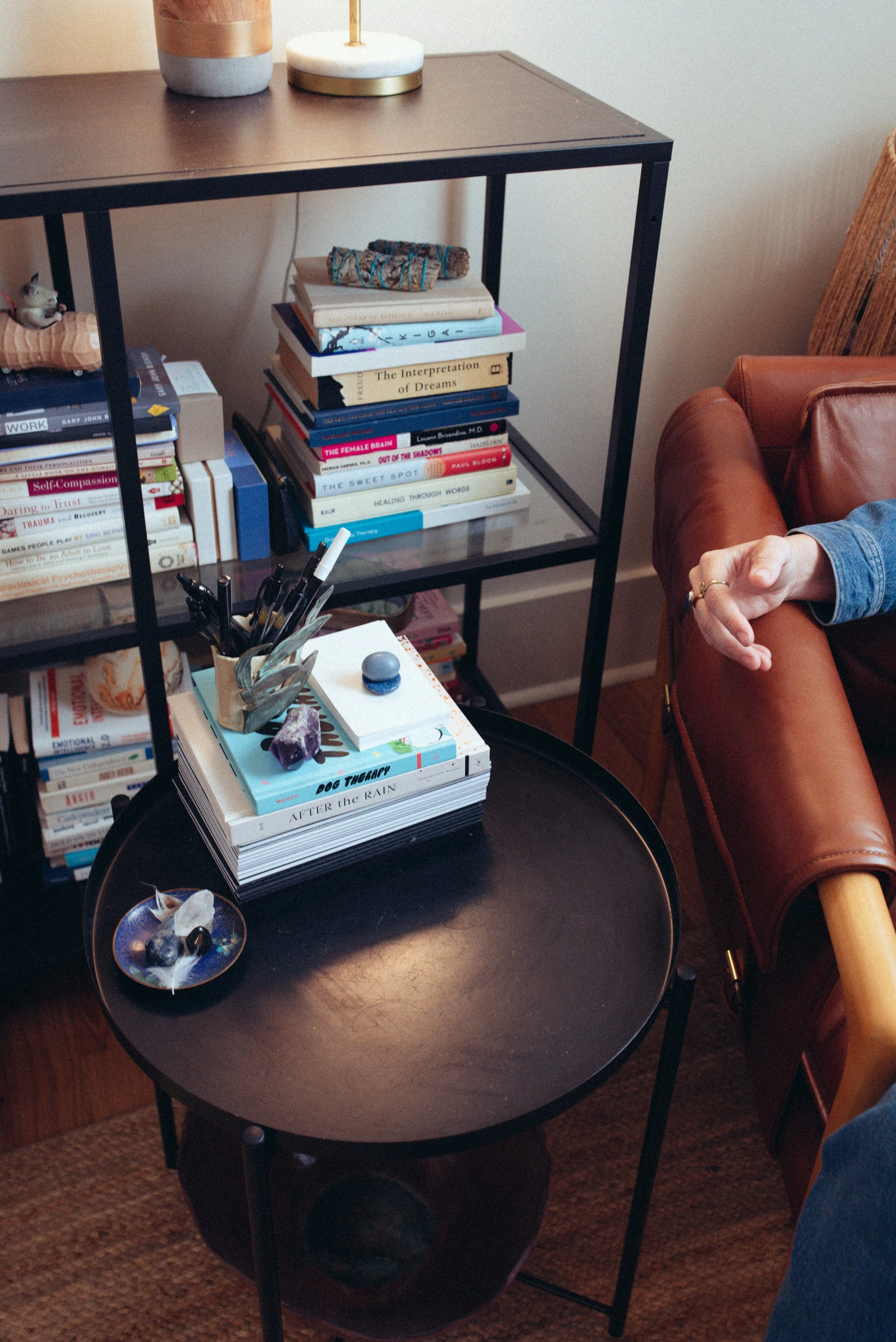 A black round side table with a stack of books and crystals on top, a small glass dish with stones, nearby a brown leather armchair with a person's arm resting on it, and a black metal bookshelf filled with more books and decorations behind.