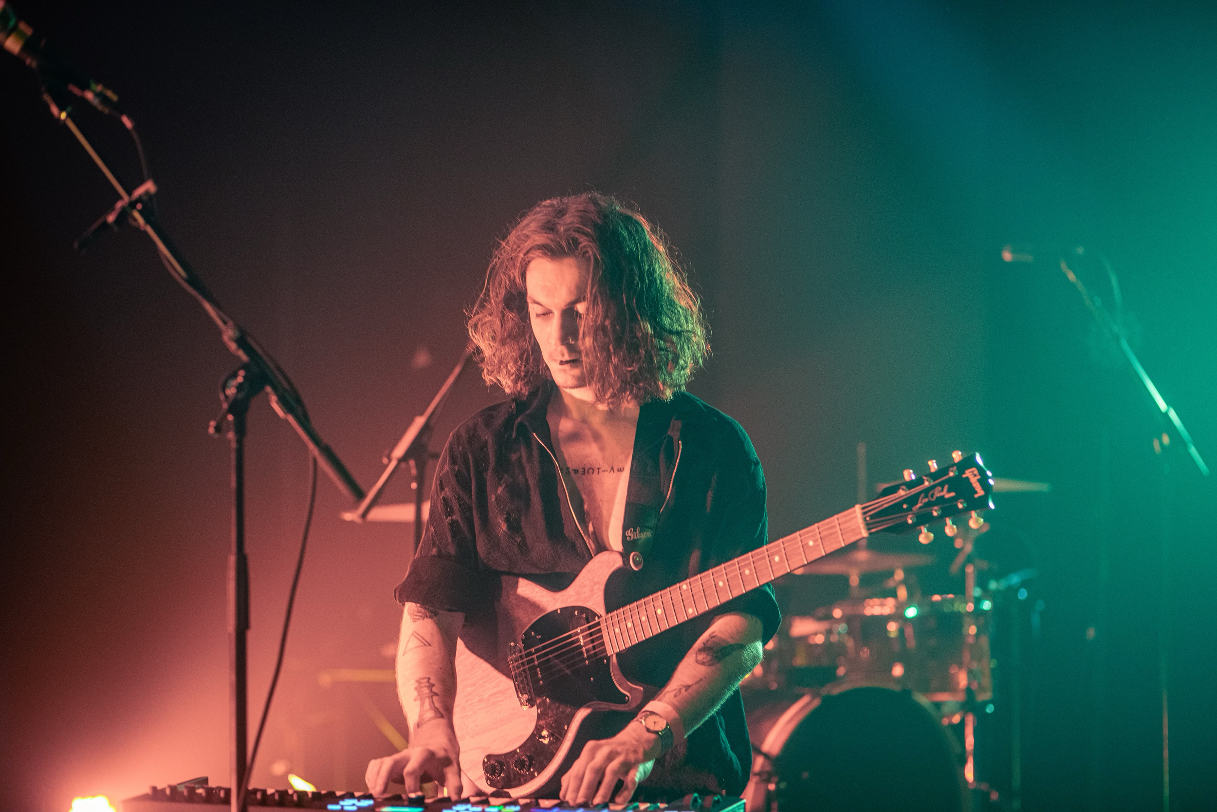 Musicien sur scène jouant de la guitare électrique et utilisant un clavier, éclairé par des lumières colorées.
