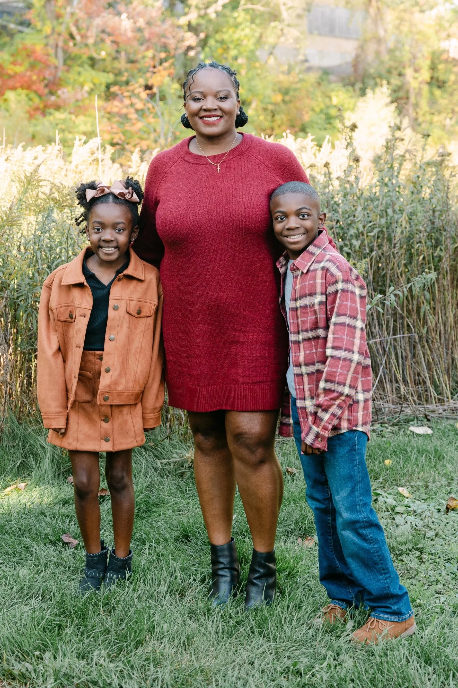 A woman with two children outdoors in a park during fall, with colorful trees in the background. The woman is wearing a red dress, and the children, a girl and a boy, are dressed in casual fall outfits. They are smiling and standing on grass.