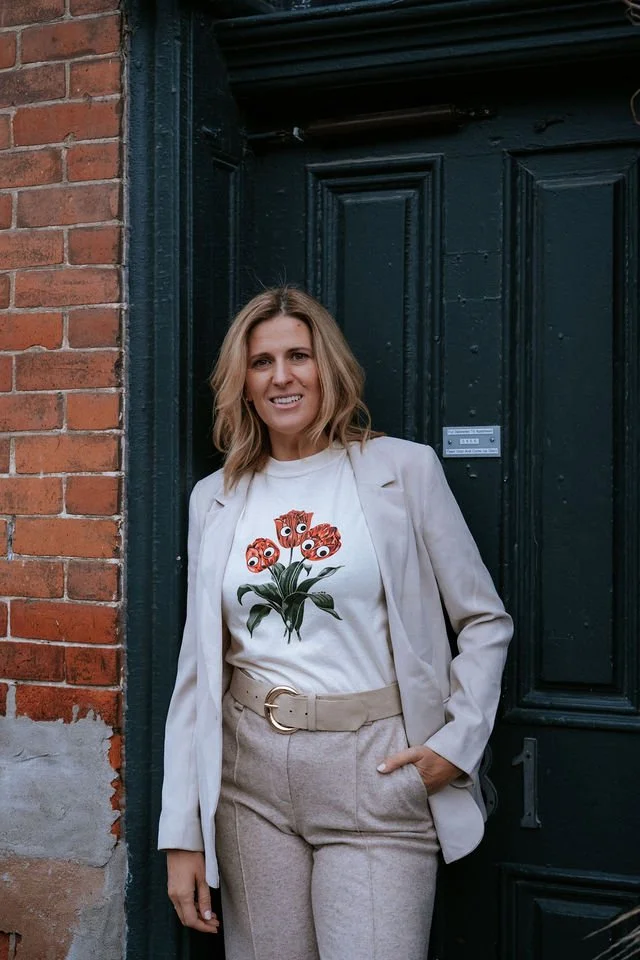 A woman with shoulder-length blond hair and a light beige blazer over a white T-shirt with a floral design, standing in front of a black door and a brick wall.