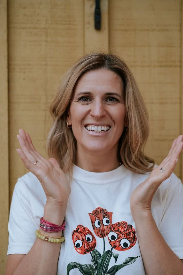 A woman smiling with hands near her face, wearing a white T-shirt with a graphic of orange tulips with cartoon eyes, standing in front of a wooden background.