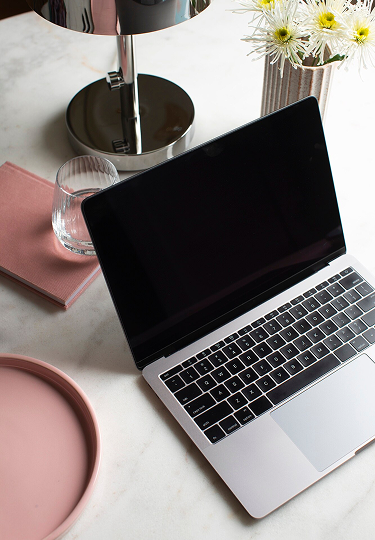 Open laptop on white table surrounded by a pink tray, glass of water, pink notebook, table lamp, and a vase with white flowers.