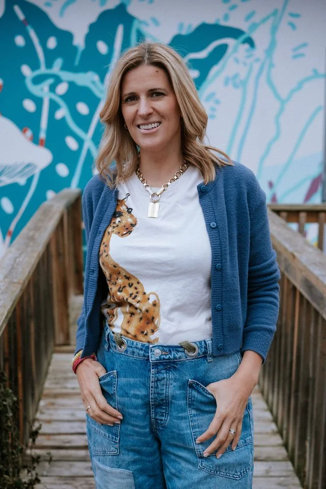 Smiling woman standing on a wooden bridge in front of a colorful mural, wearing a white t-shirt with a cheetah print, a blue cardigan, and denim jeans.