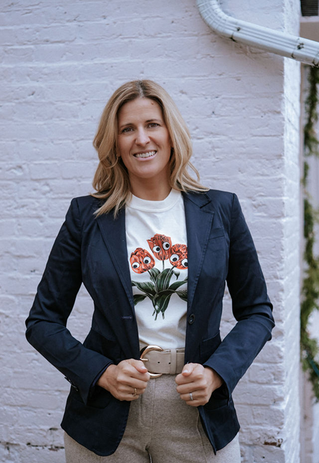 A woman with blonde hair smiling in front of a white brick wall, wearing a dark blue blazer, a white t-shirt with cartoonish flowers, and beige pants with a belt.