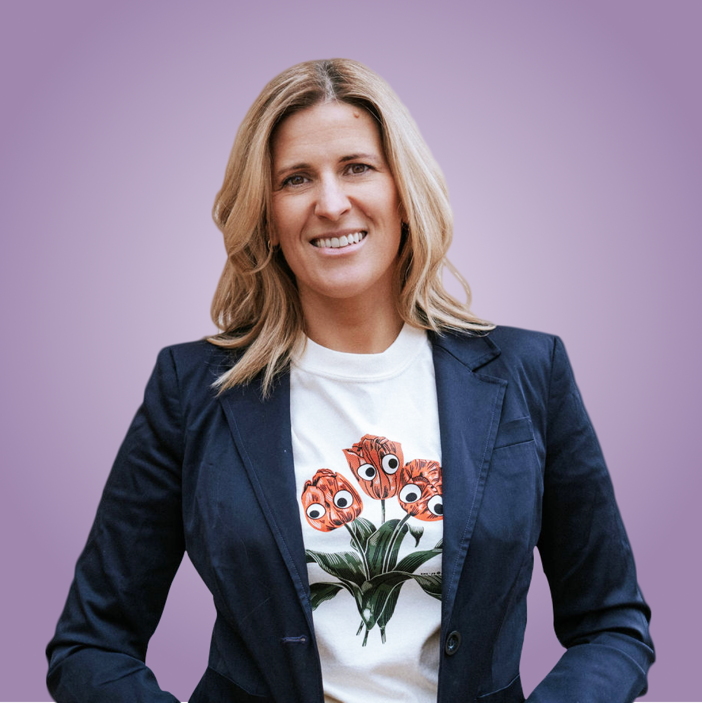 A woman with shoulder-length blond hair, wearing a dark blazer and a white t-shirt with a floral design of orange flowers with cartoonish eyes, smiling against a light purple background.