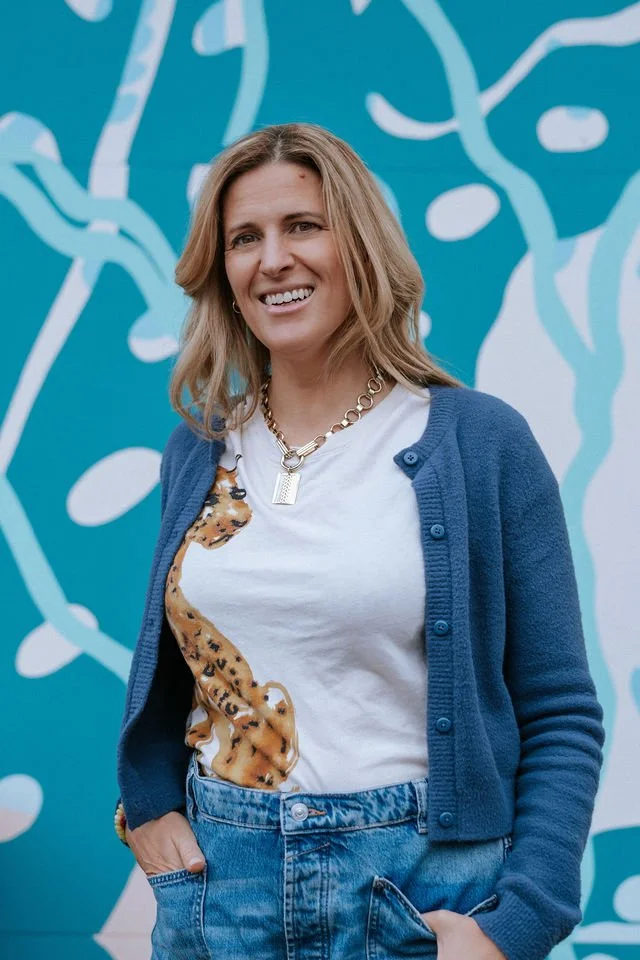 A woman with shoulder-length blonde hair, smiling, wearing a white T-shirt with a cheetah print graphic, a blue cardigan, and blue jeans, standing in front of a colorful abstract mural with blue, white, and teal shapes.