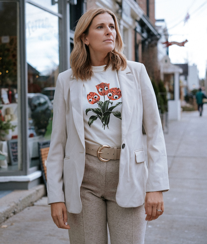 A woman with shoulder-length light brown hair wearing a white blazer, graphic t-shirt with orange flowers and eyes, beige belt and gray pants, walking on a sidewalk in a small town or shopping district.