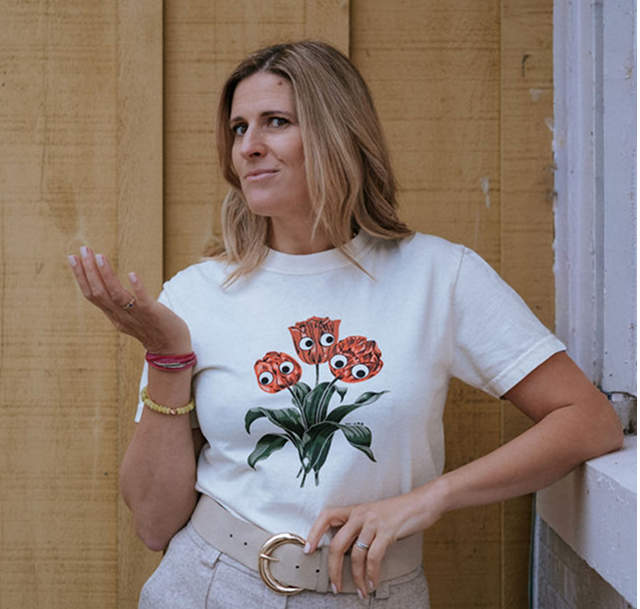 Woman with shoulder-length blonde hair wearing a white t-shirt with a graphic of orange flowers with cartoon eyes, beige pants, and a beige belt, standing against a wooden wall and a white-painted wall.