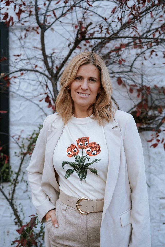 A woman with shoulder-length blonde hair smiling, wearing a white blazer, a graphic t-shirt with cartoon-like flowers with faces, and beige high-waisted pants with a belt, standing outdoors in front of a tree with reddish leaves.