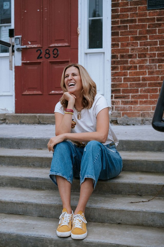 A woman sitting on the front steps of a brick house, smiling and laughing, with a red front door and a window behind her.
