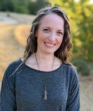 A woman with wavy brown hair smiling outdoors, wearing a dark gray top and a necklace with a pendant.