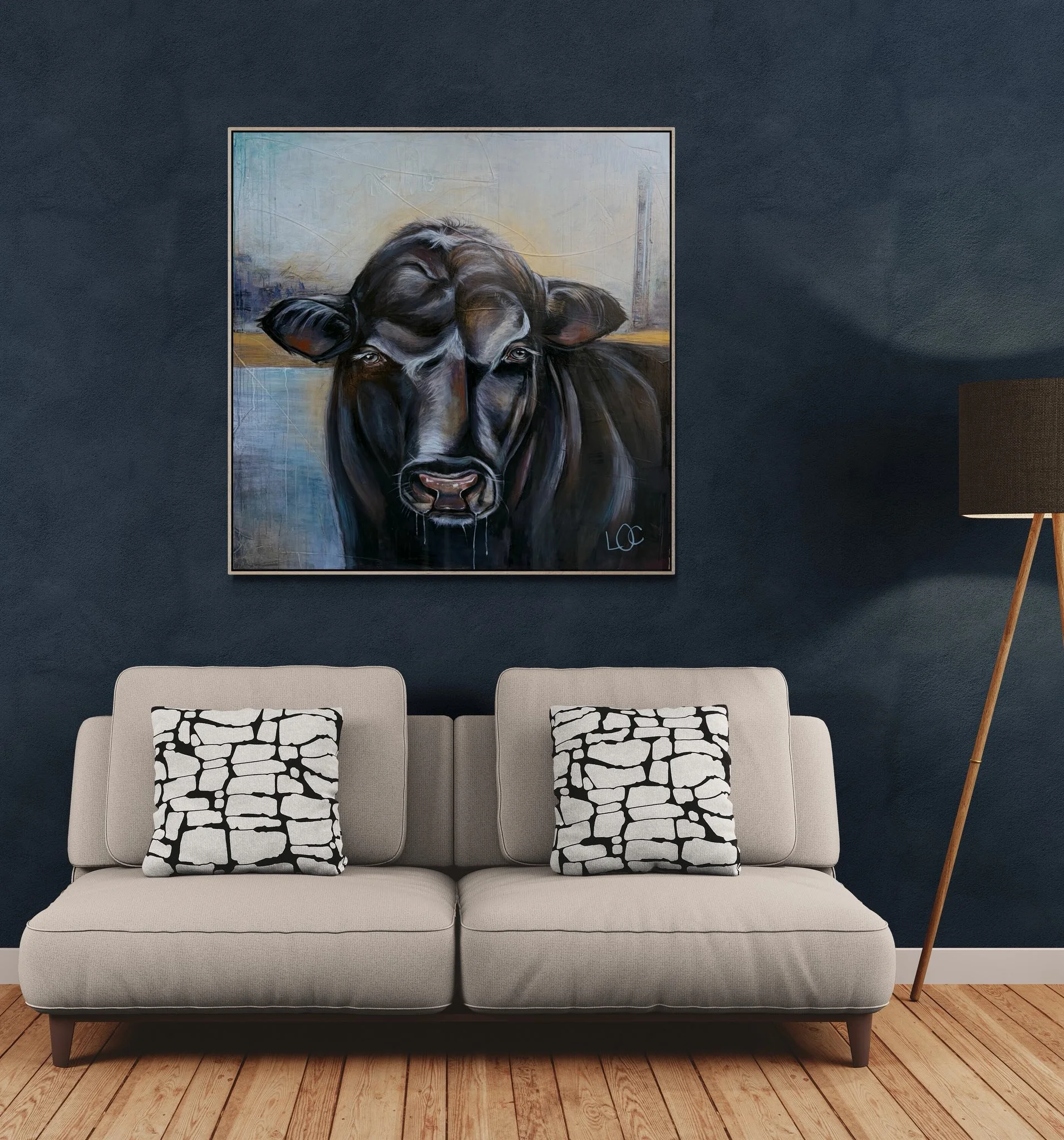 A modern beige sofa with three black and white patterned pillows, positioned on a wooden floor against a dark blue wall. A framed painting of a black cow hangs above the sofa, and a small wooden side table with a lamp is partially visible on the righ