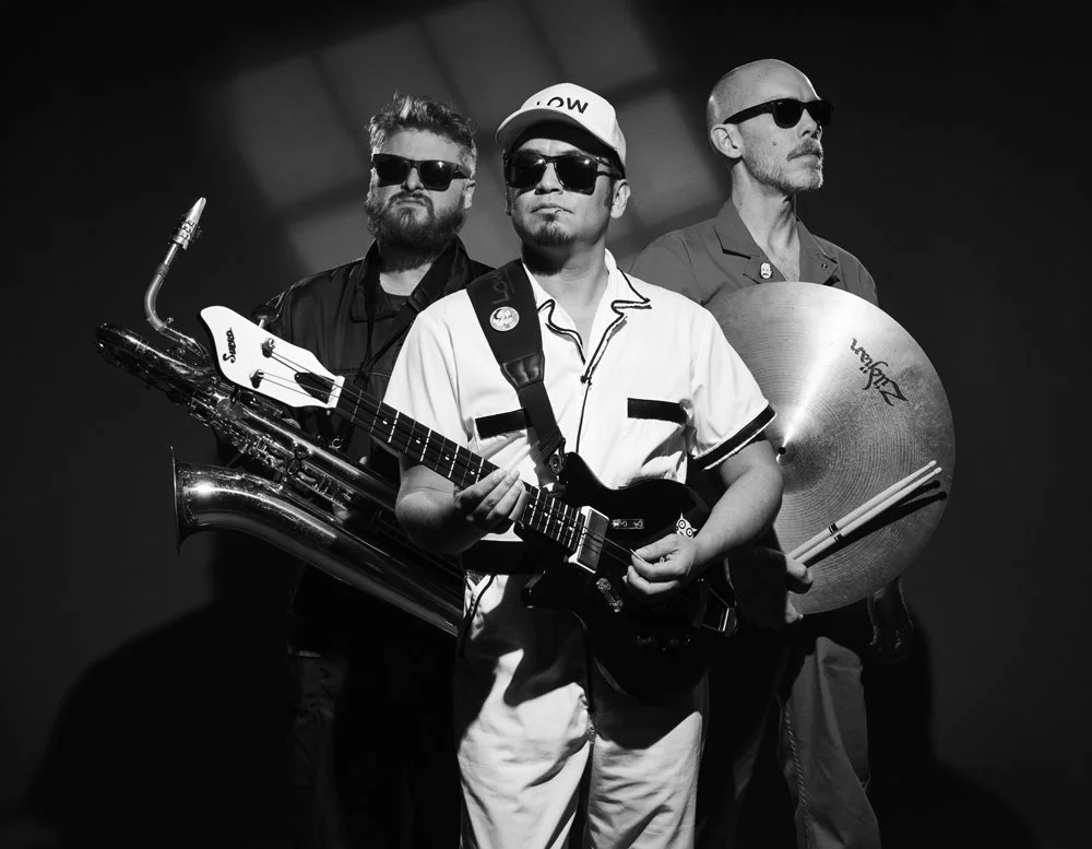 Three men with sunglasses holding musical instruments, standing against a dark background.
