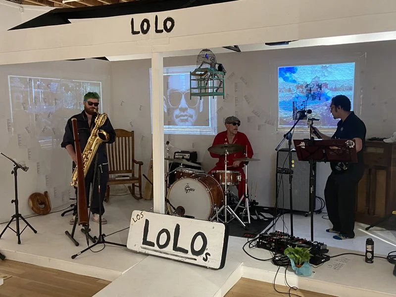 A band called LOLO performing on a small stage with a drummer, a saxophonist, and a keyboard player. Behind them are two screens, one showing a person's face and the other a boat on water. The stage is decorated with a sign that says "LOLO" and a small hanging birdcage.