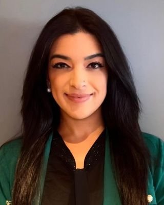 A woman with long black hair smiling, wearing a black top and a green jacket, against a neutral background.