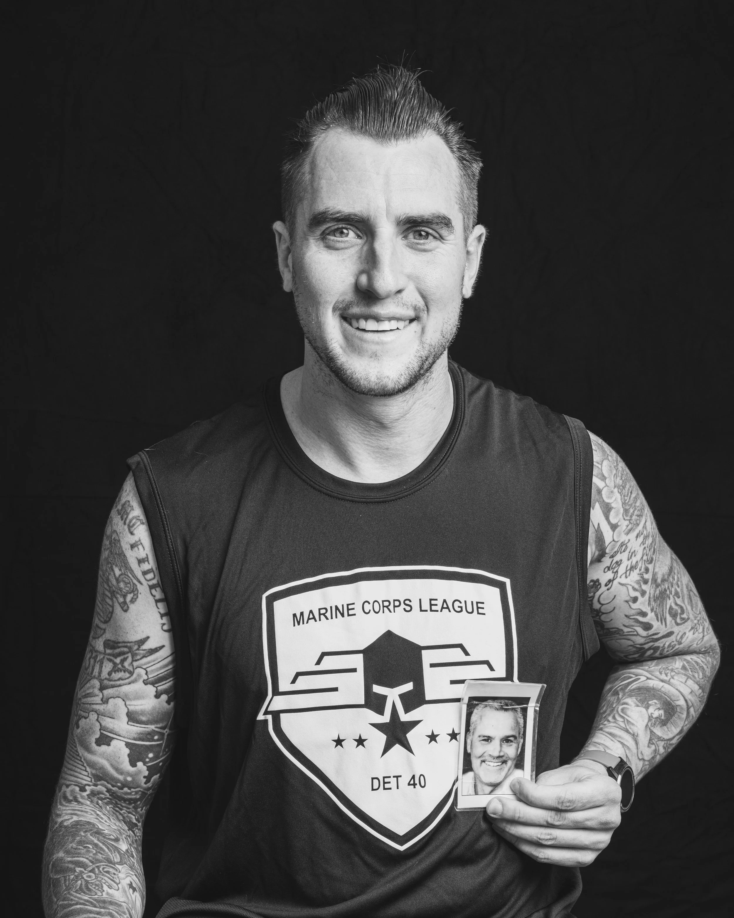 A smiling man with tattoos on his arms, wearing a sleeveless shirt with a Marine Corps League logo, holding a small portrait of himself, against a dark background.